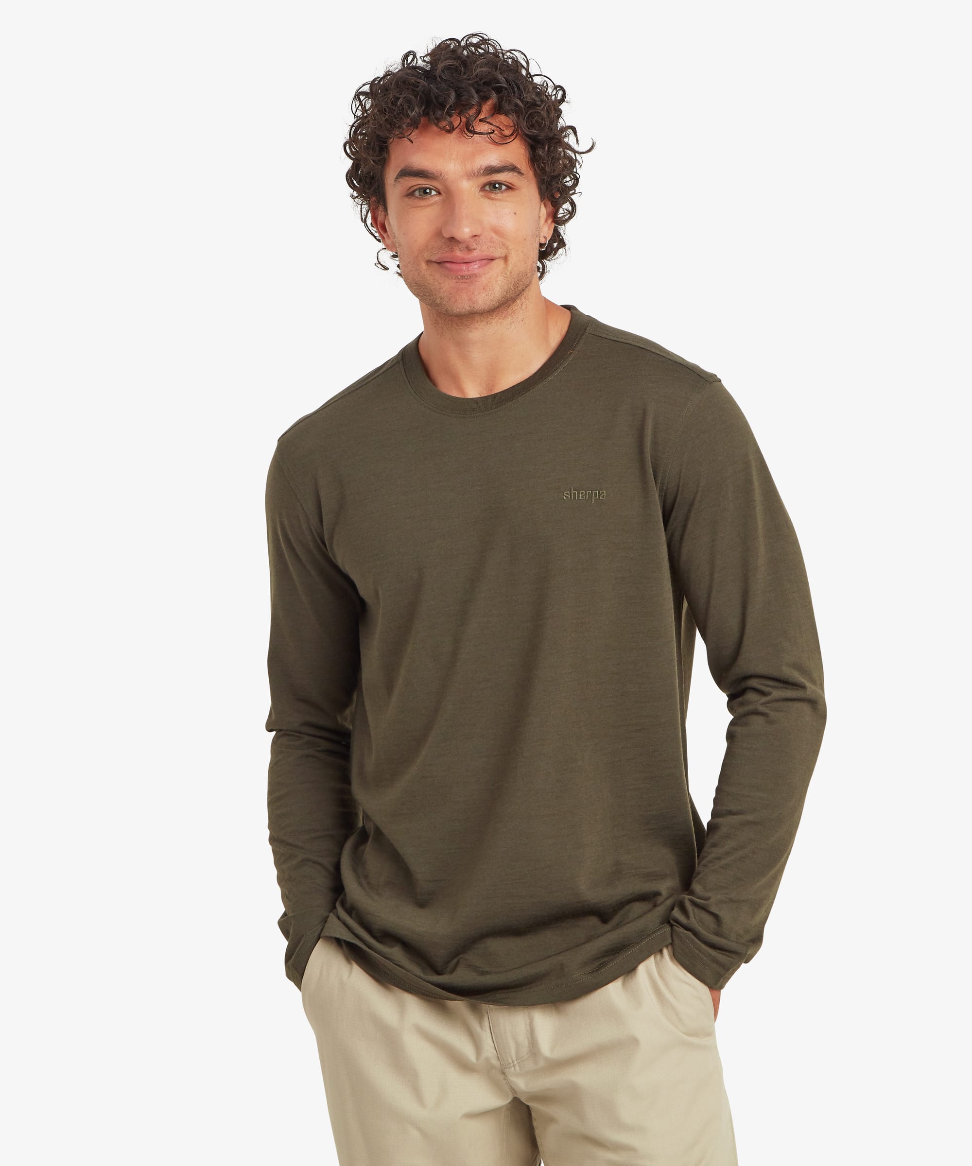 Man wearing an olive green long-sleeve merino wool tee with a classic crew neck and regular fit. He stands casually with both hands in his pockets, paired with light beige pants. A small tonal logo is embroidered on the chest.