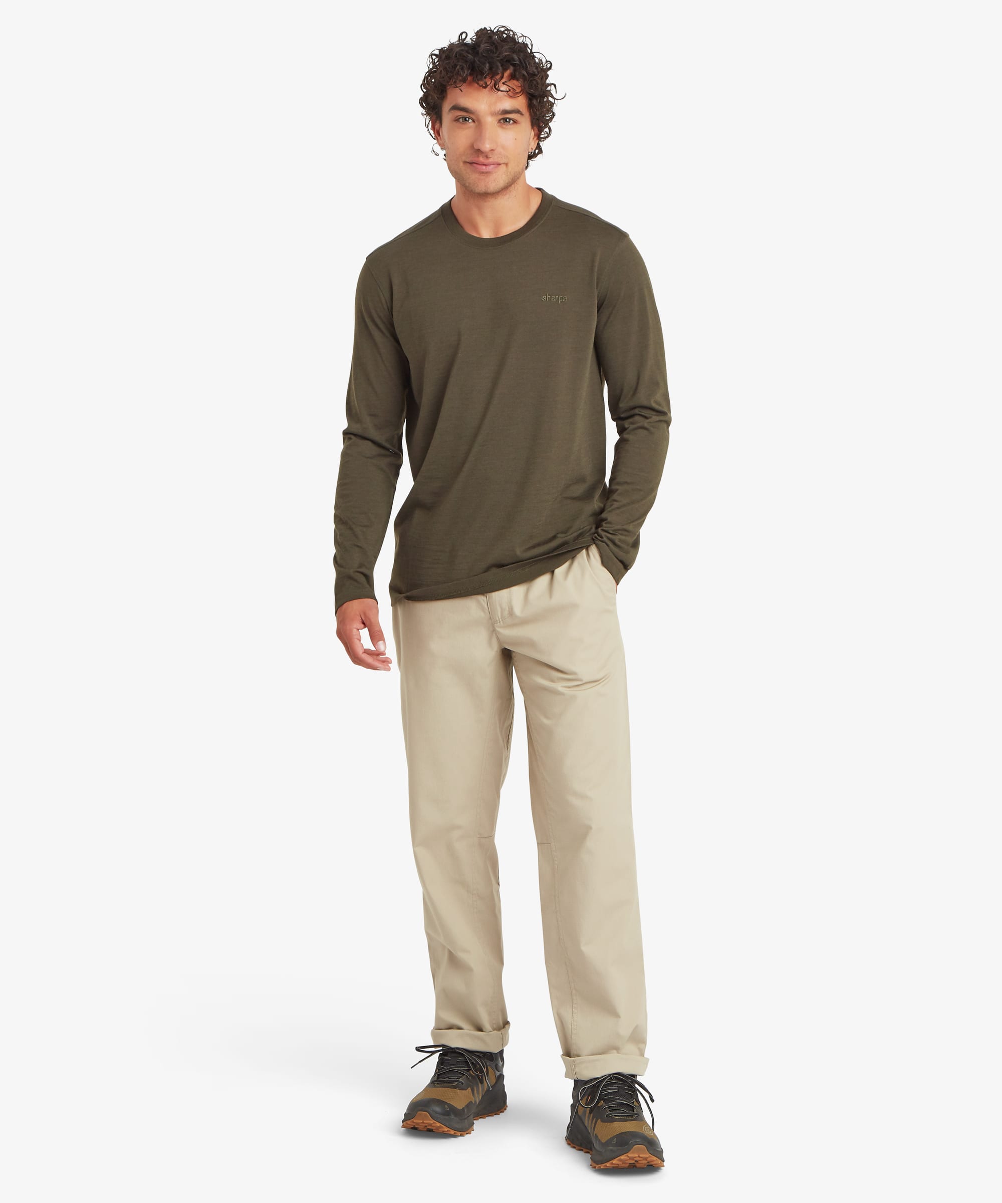 Full-body front view of a man wearing an olive green long-sleeve merino wool tee. The lightweight top features a clean crew neckline and subtle chest logo, paired with beige pants and hiking shoes.