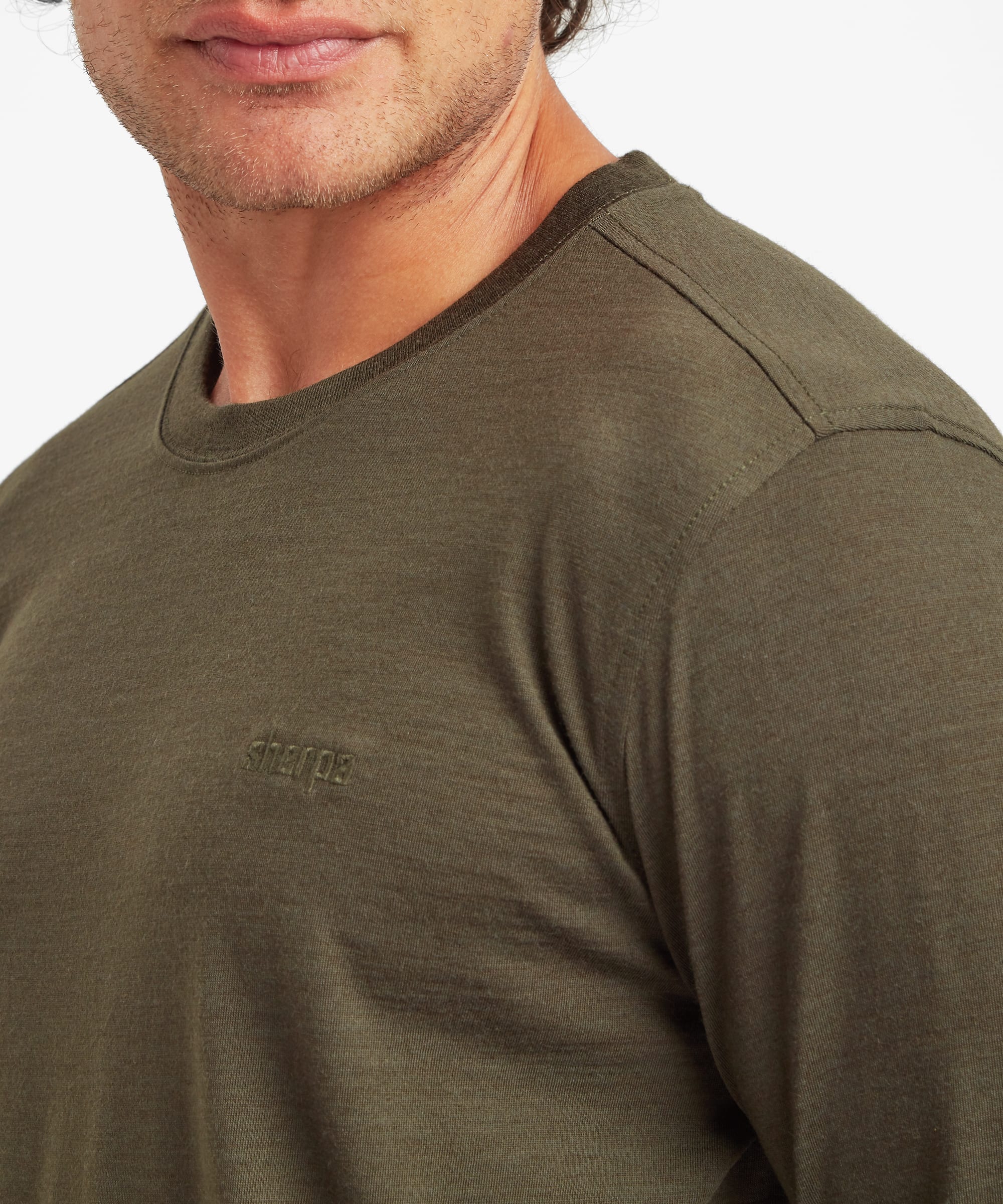Detailed close-up of the olive green merino wool tee highlighting the crew neckline, fine knit texture, shoulder seam, and subtle tonal logo embroidered on the left chest.