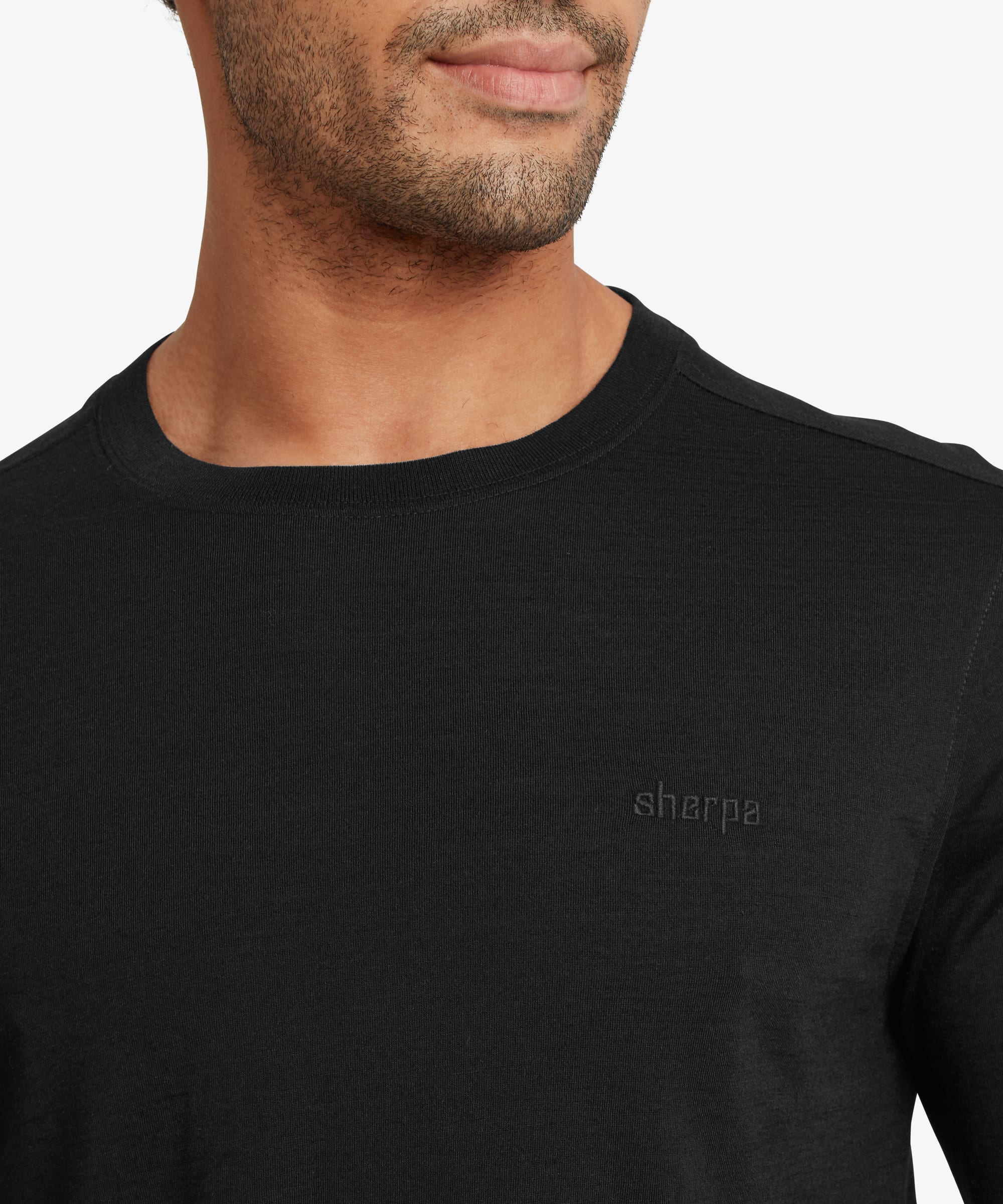 Close-up of the black merino wool long-sleeve tee showing the crew neckline, fine knit fabric, and subtle tonal embroidered logo on the left chest. The lightweight texture and clean finish are highlighted.