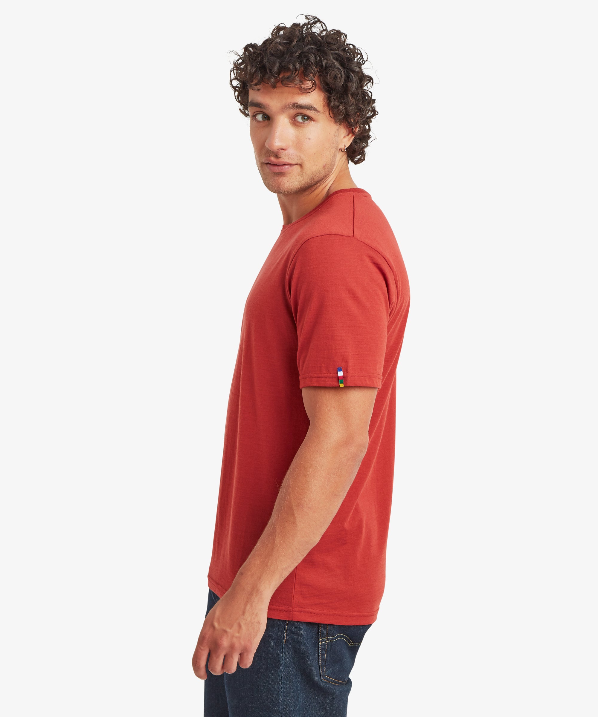 A side profile of a rust‑red Merino wool short-sleeve T‑shirt paired with dark denim jeans, with a small multicolored woven label visible on the sleeve.