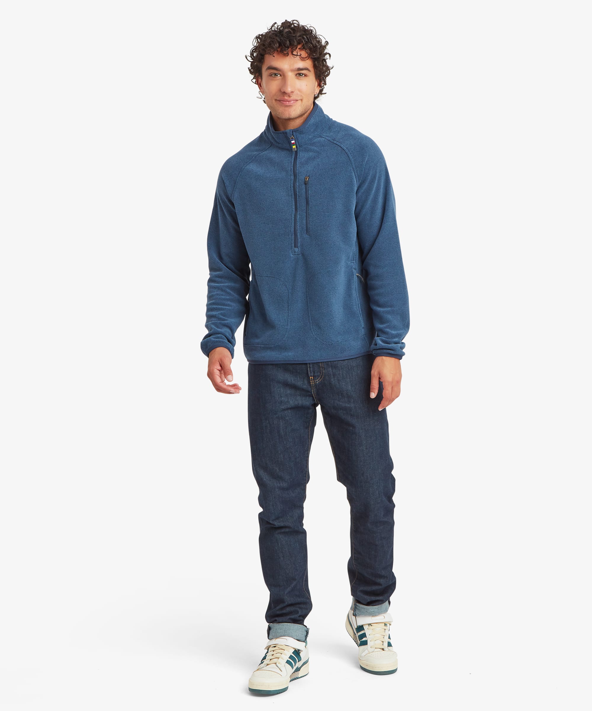 Full-body front view of the blue Rolpa 1/2-zip fleece. The regular-fit fleece features a high collar, chest zip pocket, and zippered hand pockets, paired with dark pants and light-colored sneakers.