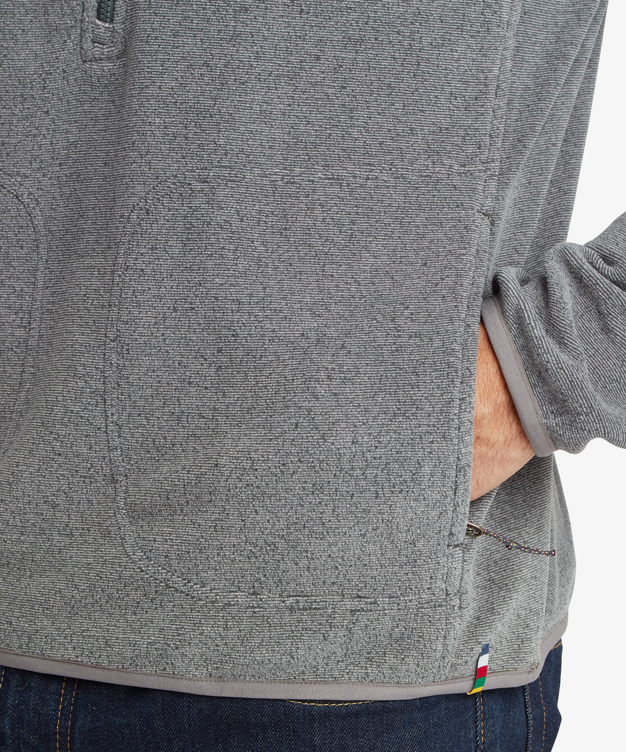 Detailed close-up of the lower section of the grey fleece showing the zip hand pocket, textured fleece fabric, and small multicolor accent tab near the hem.