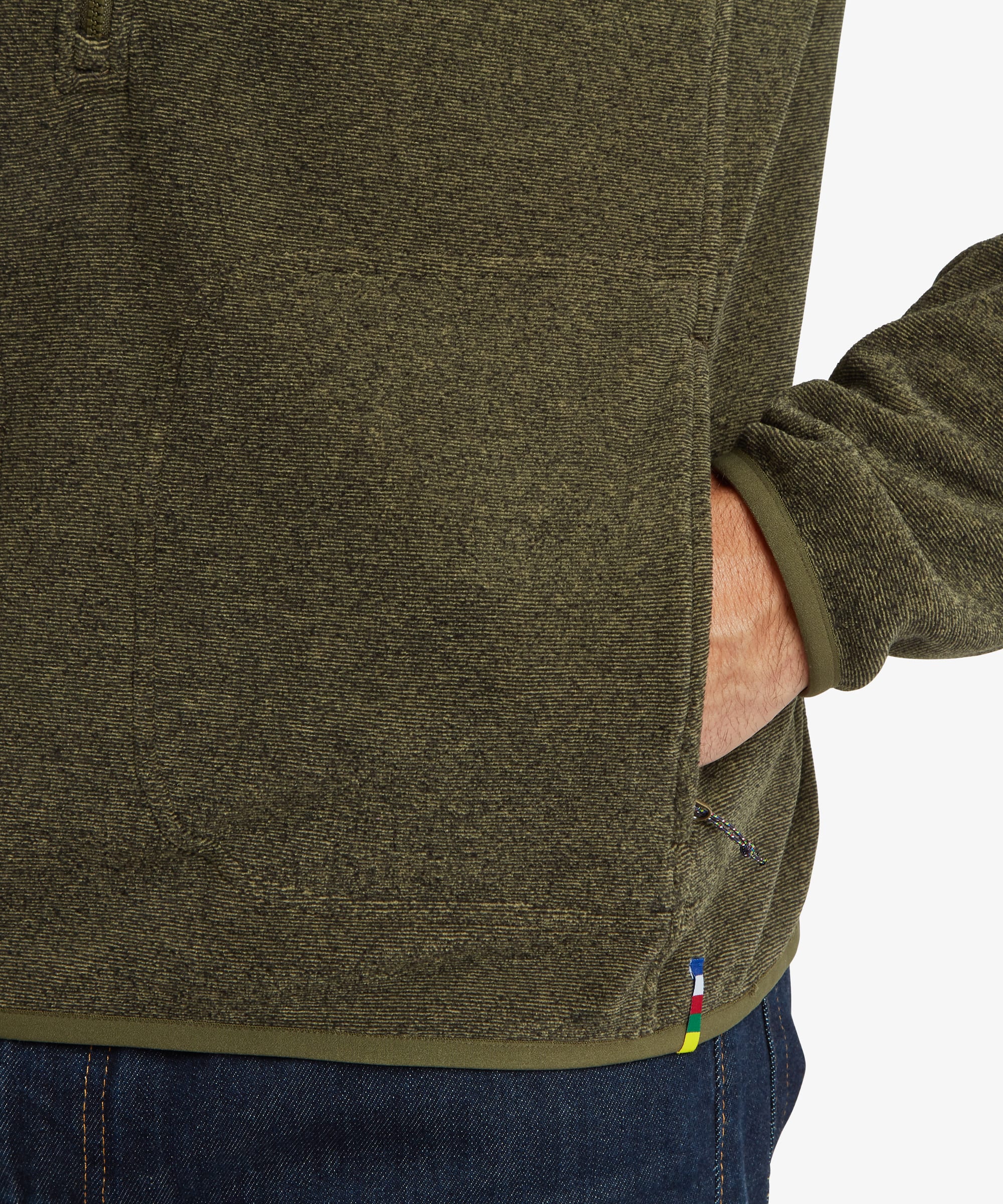 Close-up of the lower side pocket area of an olive green fleece. A hand rests inside the zippered pocket, highlighting the practical storage feature and subtle trim detail.