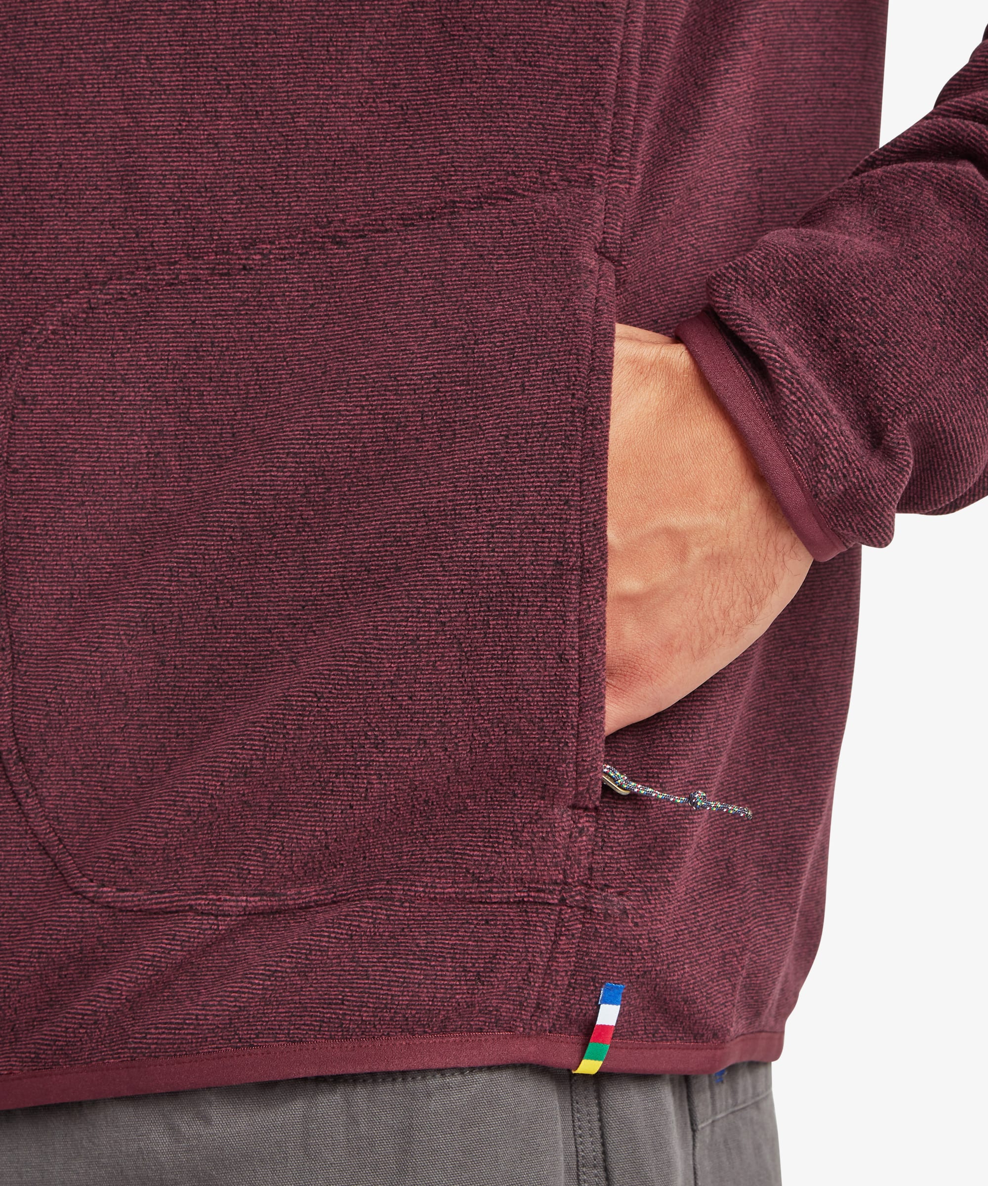 Detailed close-up of the burgundy fleece jacket showing the zip hand pocket, fleece texture, and multicolor accent tab near the hem. A hand is placed inside the pocket to demonstrate storage.