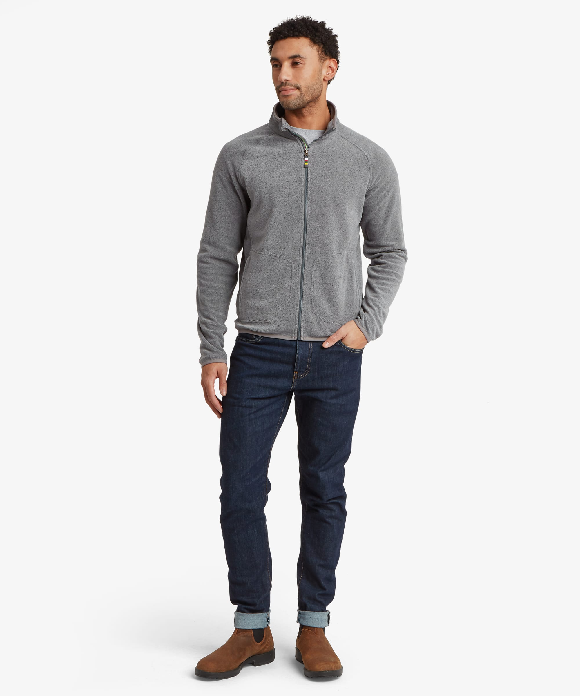 A man modeling a gray full fleece jacket, dark blue jeans, and brown boots against a light gray studio background.The fleece features grey binding on the hem and cuff and a prayer flag zip puller. The man is looking slightly to his right with one hand in his pocket.