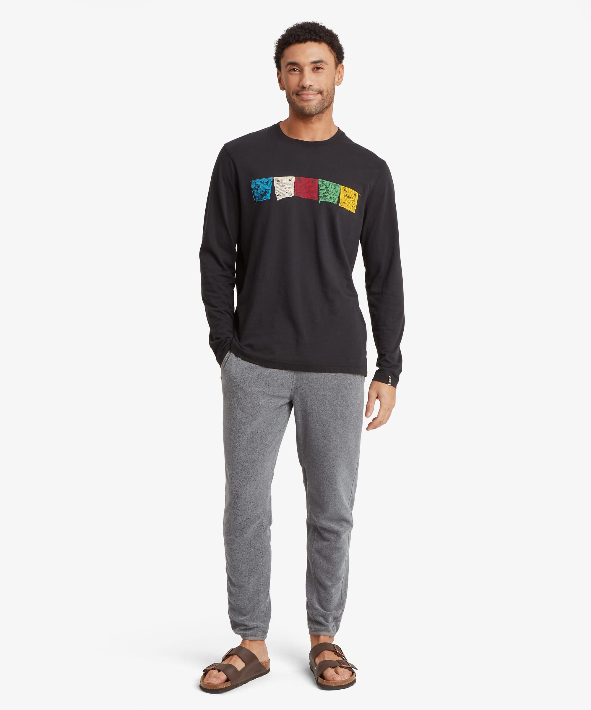 Full-body front view of a man wearing grey fleece joggers with an elastic waistband and tapered fit. The joggers feature side pockets and ribbed ankle cuffs, paired with a graphic sweatshirt and sandals.