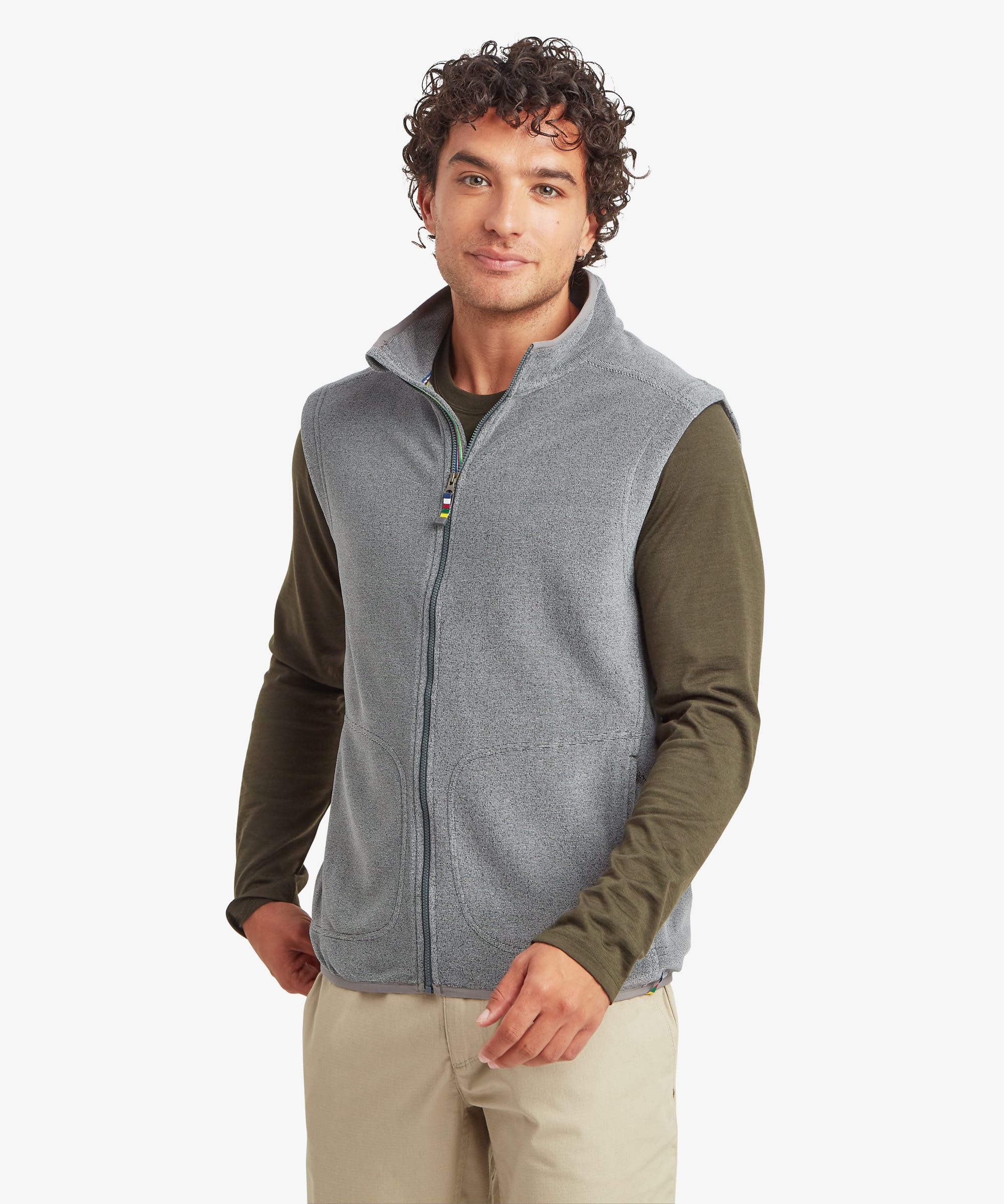Man wearing a grey fleece vest with a stand collar and full-length front zipper. The sleeveless vest features zippered hand pockets and contrast olive sleeves from the layered shirt underneath. He stands smiling with one hand in a pocket, paired with beige pants.