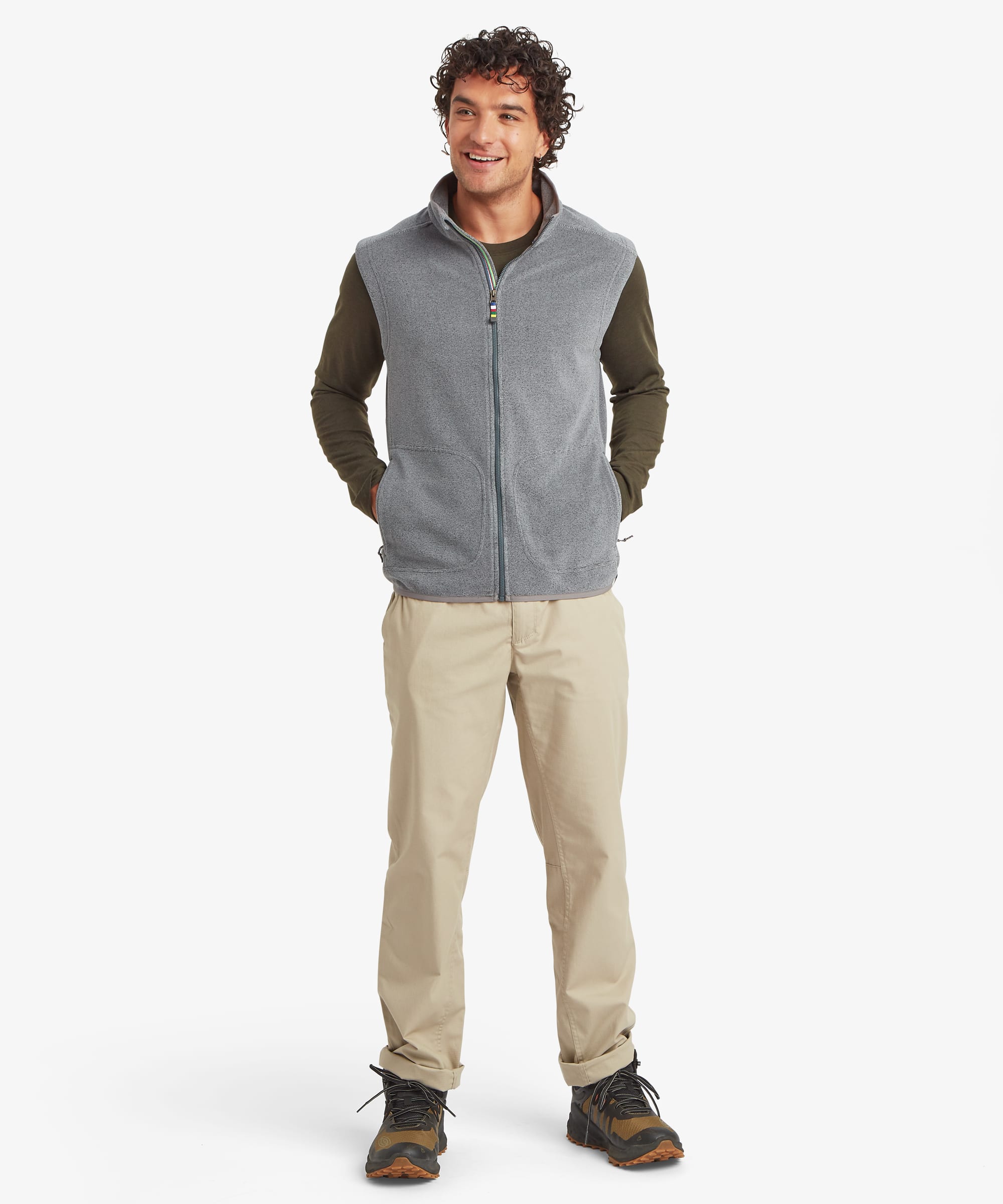 Full-body front view of a man wearing a light grey fleece vest with a stand collar and full-length front zipper. The sleeveless vest features zippered hand pockets and multicolor trim accents at the zipper. Styled over an olive long-sleeve shirt with beige pants and hiking shoes.