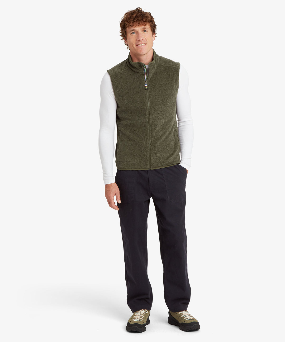 A man modeling an olive green fleece vest over a white long-sleeve shirt, dark pants.