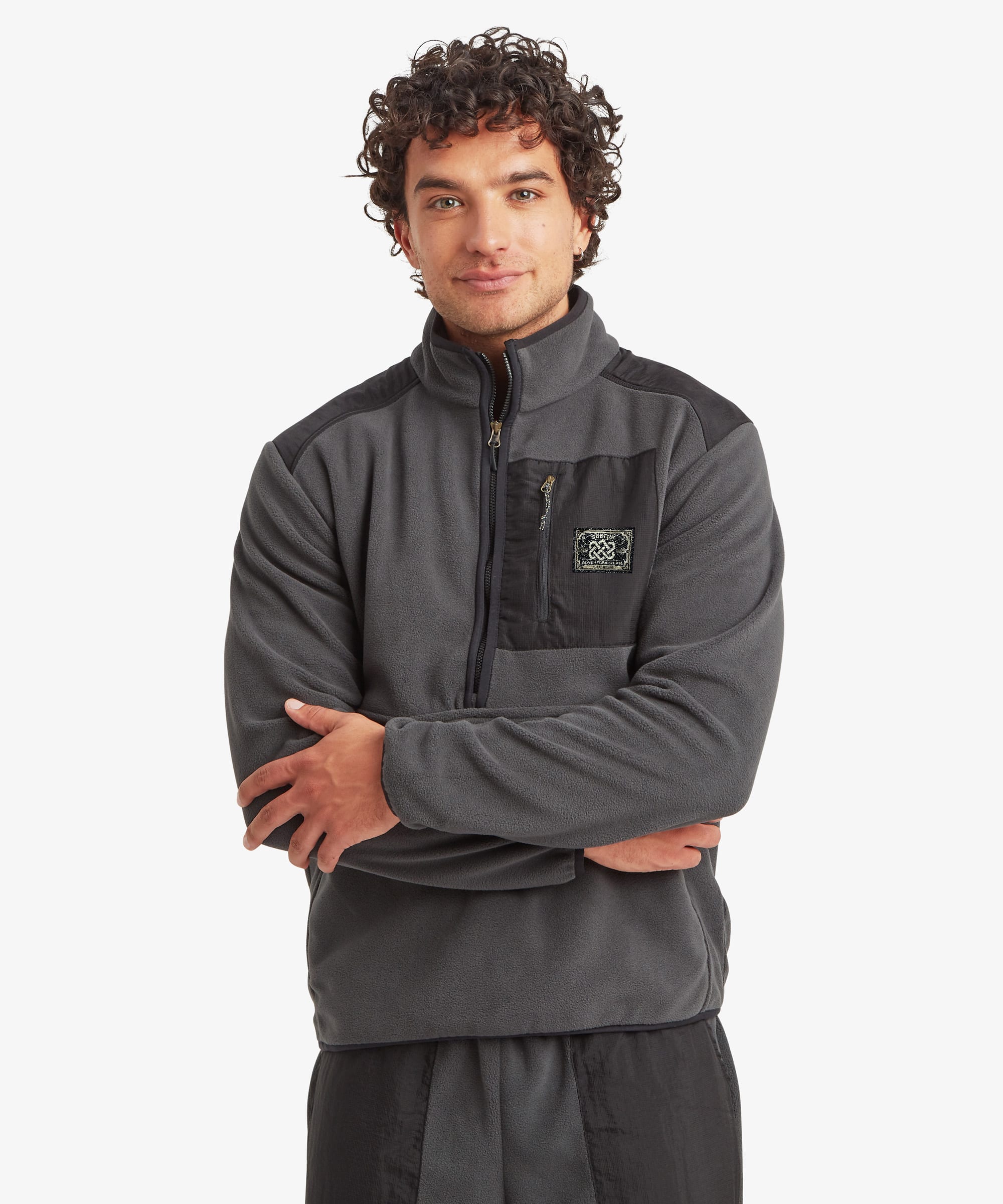 A man wearing a dark gray half-zip fleece pullover with contrasting black shoulder panels and a small chest Sherpa Adventure Gear logo patch on the pocket, photographed against a light gray studio background. The zip features black trim and a multi-coloured string zip puller.