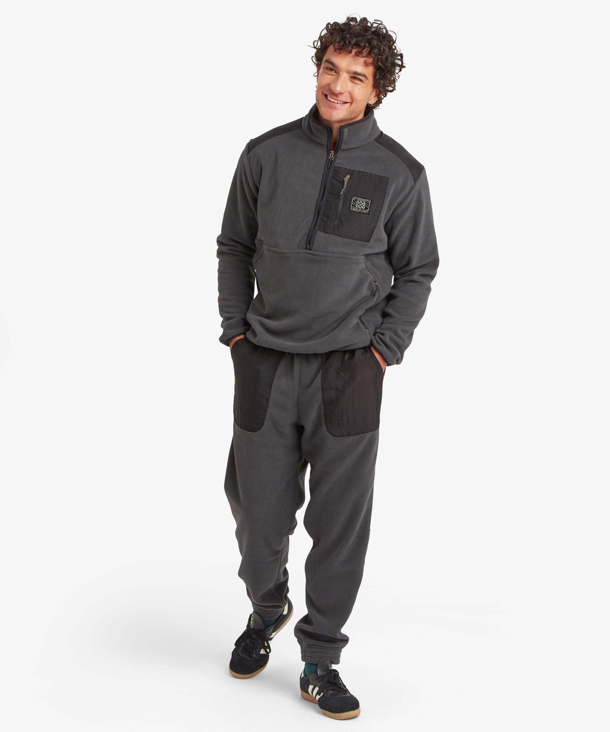 A man modeling a dark gray fleece pullover and matching jogger pants with black contrast panels, standing against a light gray studio background.The Fleece zip pullover features a Sherpa Adventure Gear patch on the chest pocket.