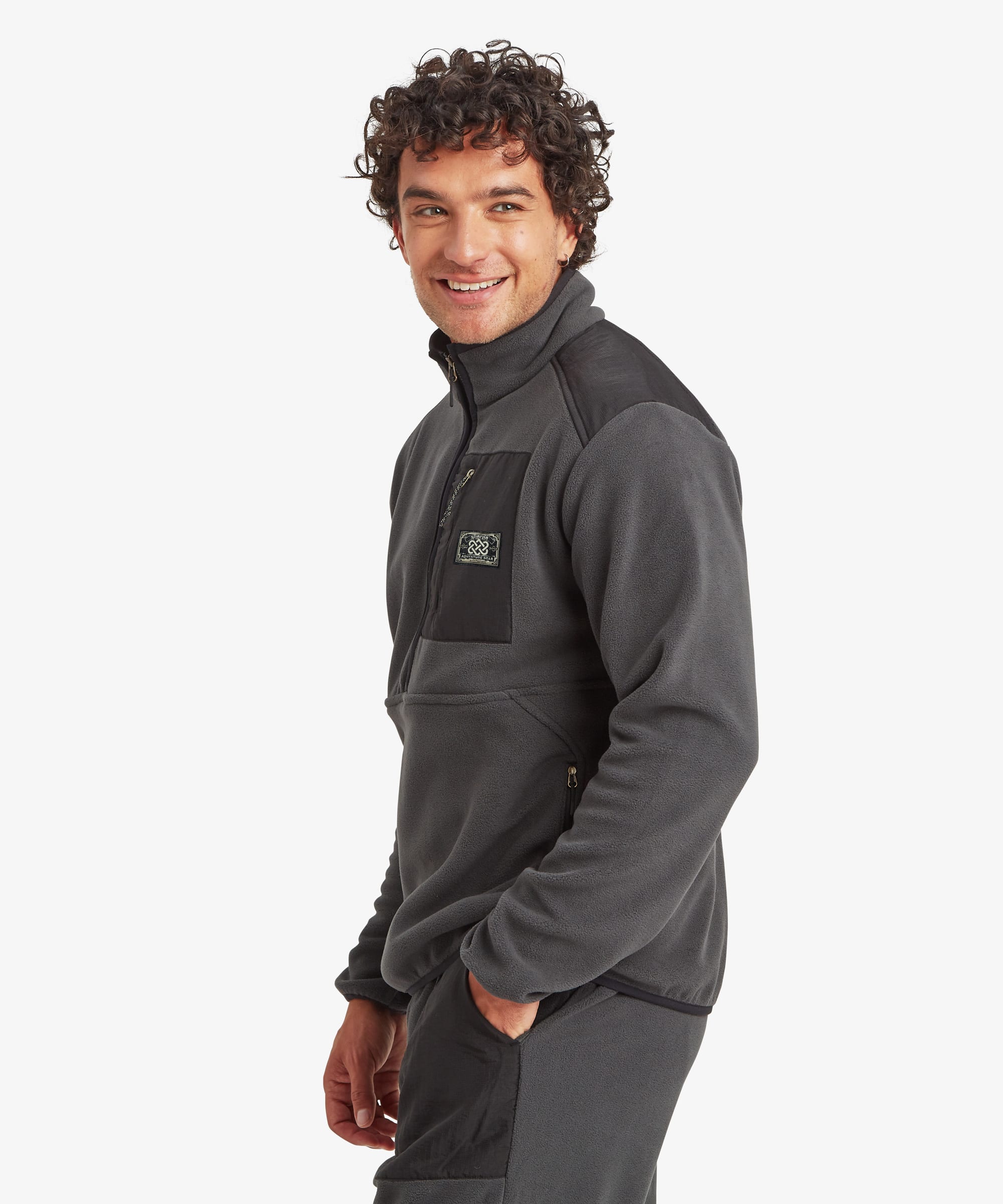 A man wearing a dark gray fleece half-zip pullover with a black chest pocket and a small Sherpa Adventure Gear logo patch and matching jogging pants, photographed against a light gray studio background. The man is looking over his shoulder with one hand in his pocket.
