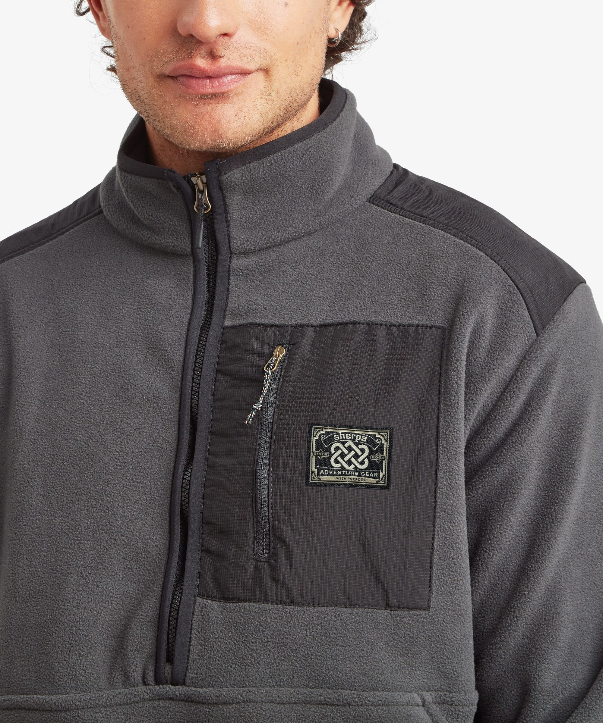 A close-up of a man wearing a dark gray fleece half-zip jacket with black shoulder panels and a black chest pocket featuring a small Sherpa Adventure Gear patch. The fleece features multi-colored zip pullers.