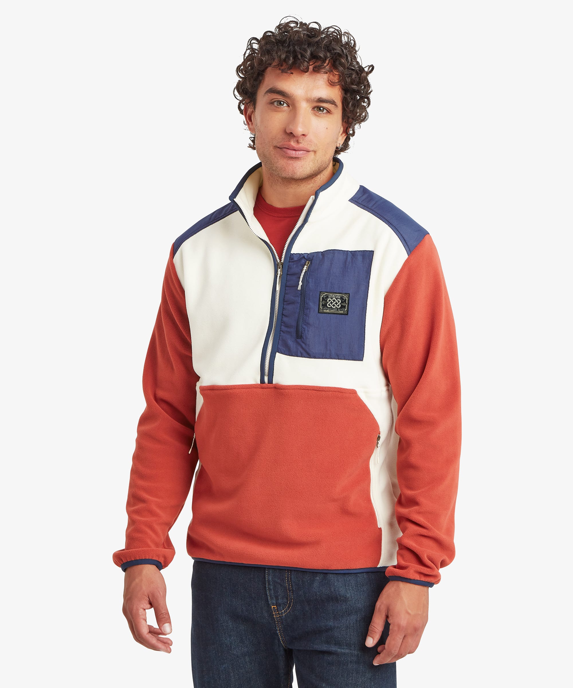 A man wearing a color-blocked half-zip fleece pullover by Sherpa Adventure Gear, featuring a rust-orange body, white chest panels, and navy blue shoulder and pocket details, paired with dark blue jeans against a light gray background.