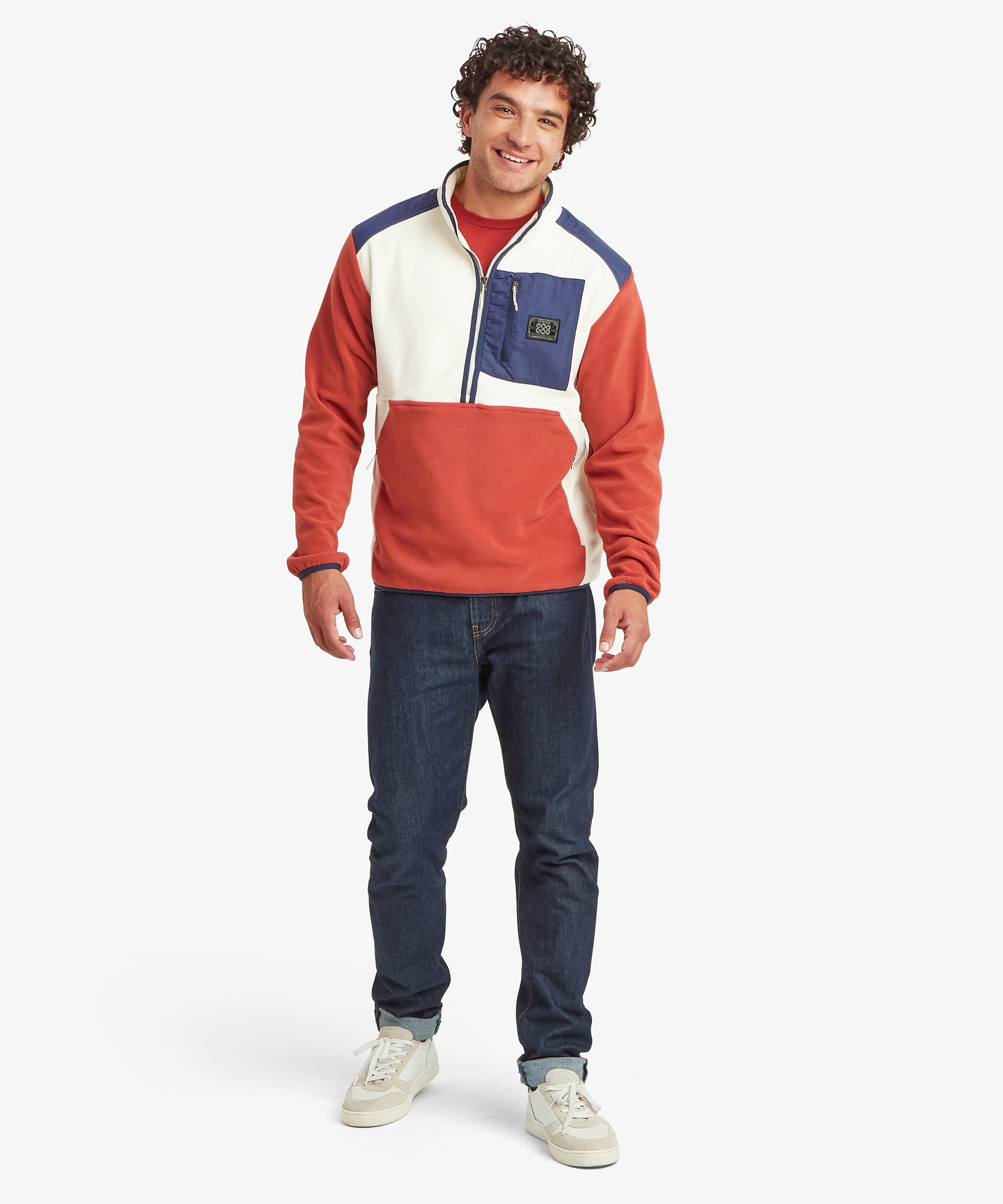 A man wearing a color-blocked fleece pullover jacket in red, white, and blue panels, paired with dark blue jeans and white sneakers, standing against a light gray studio background.
