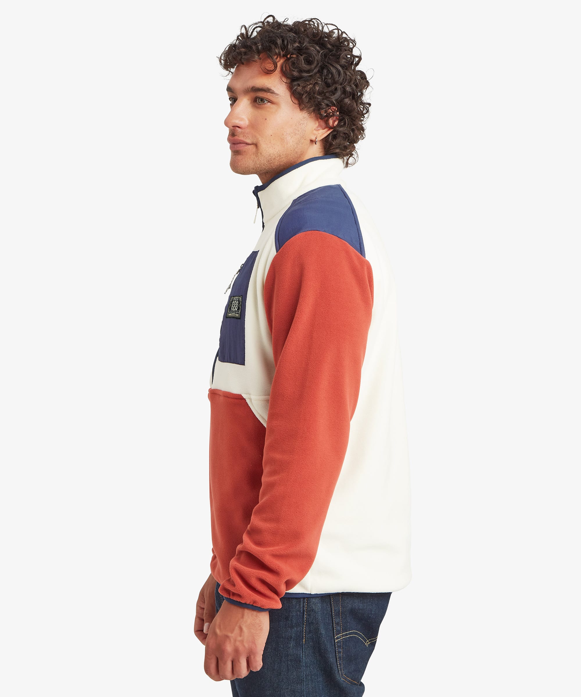 A man wearing a color-blocked half-zip fleece pullover sweater in red, navy blue, and off-white, shown in profile against a light gray background. The fleece features blue binding on the cuffs, hem and collar and a black Sherpa Adventure Gear patch on the chest.