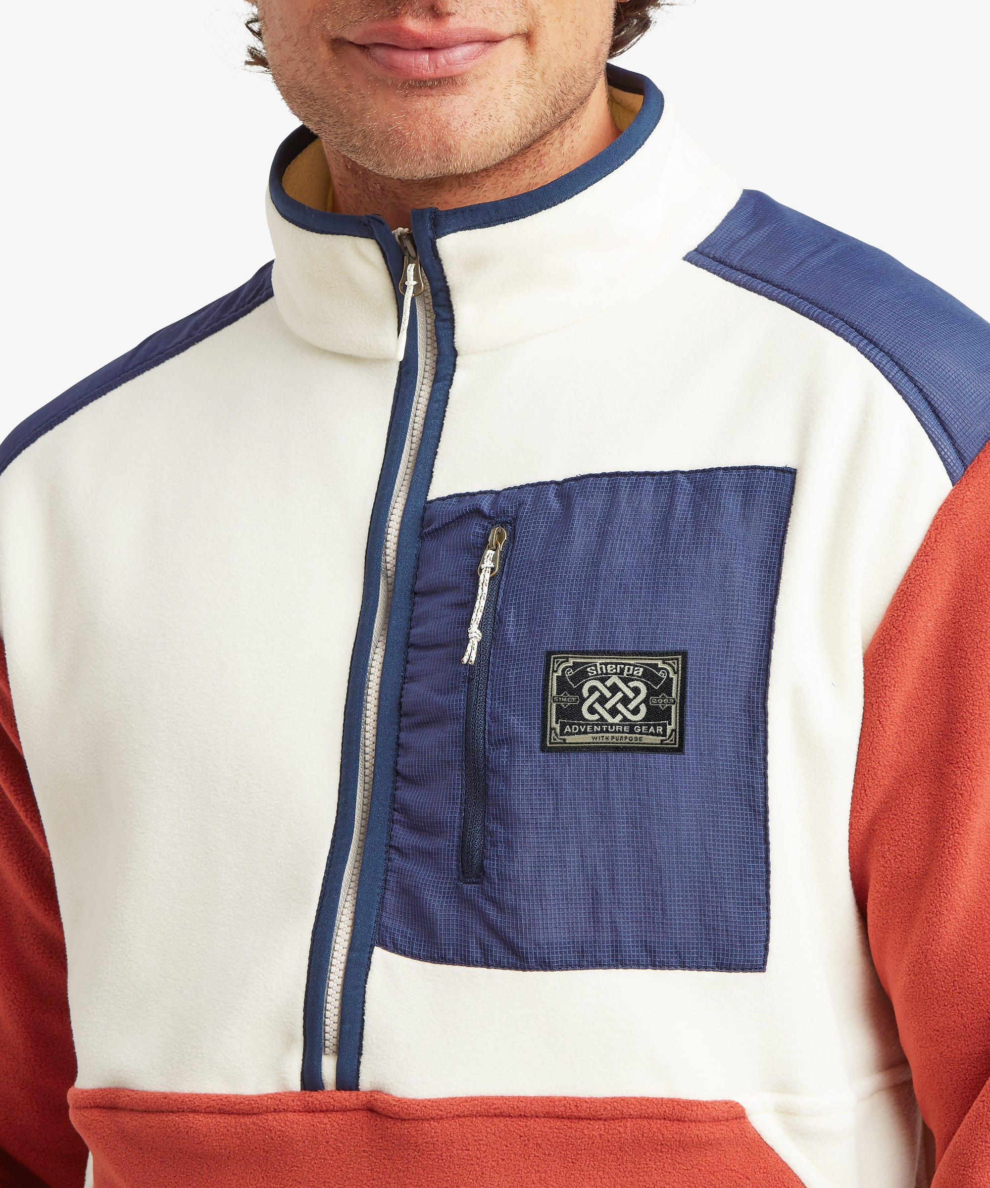 A close-up of a person wearing a color-block quarter-zip fleece jacket. The jacket features an off-white main body, a navy blue chest pocket with a zippered pocket and a black 'Sherpa Adventure Gear' brand patch, and rust-red sleeves and lower hem.