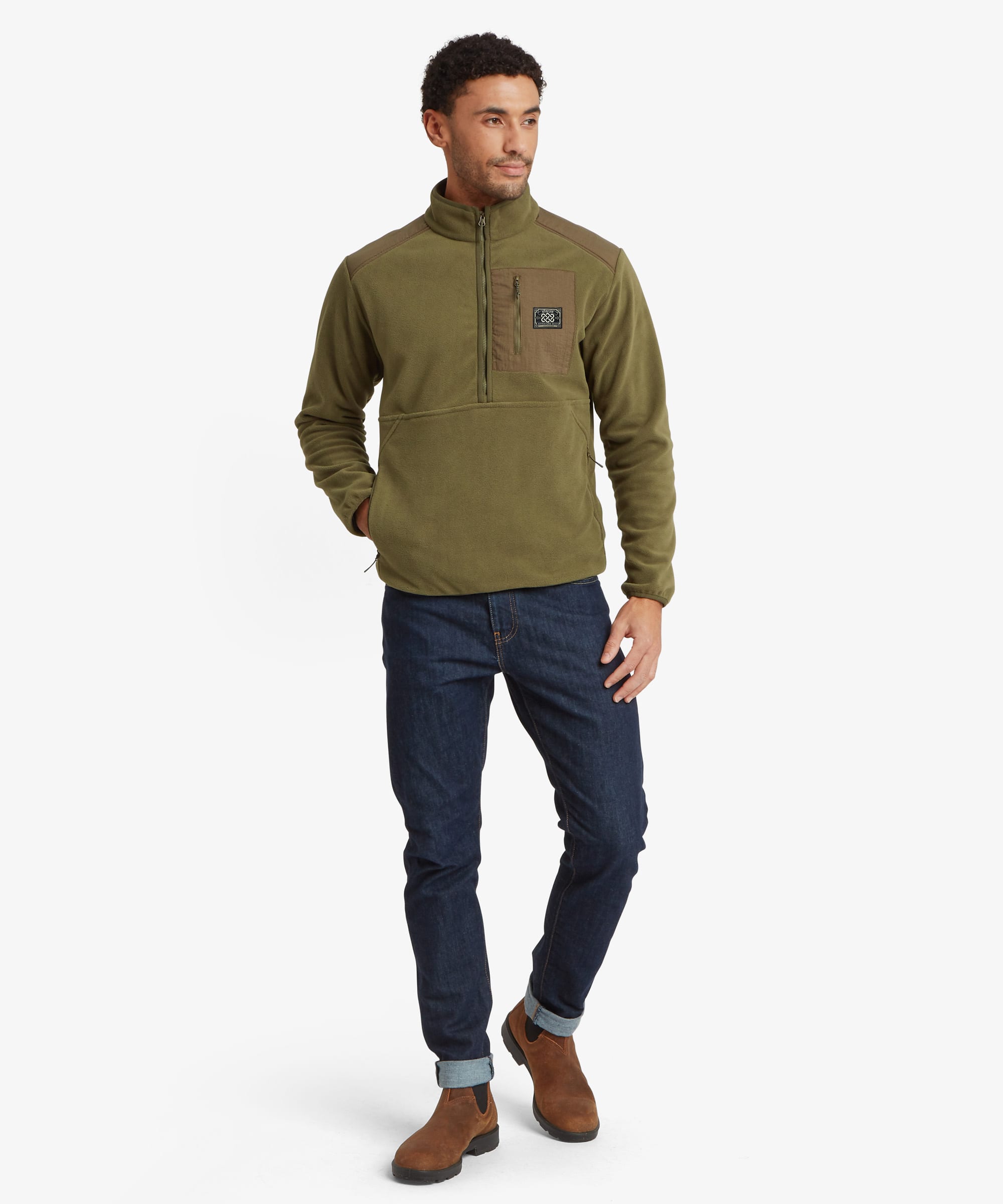 A man modeling a half-zip fleece pullover in olive green with a brown chest pocket and a small Sherpa Adventure Gear logo patch, paired with dark blue jeans and brown boots, against a light gray studio background. The model is looking to his right with his hand in the fleece pocket.