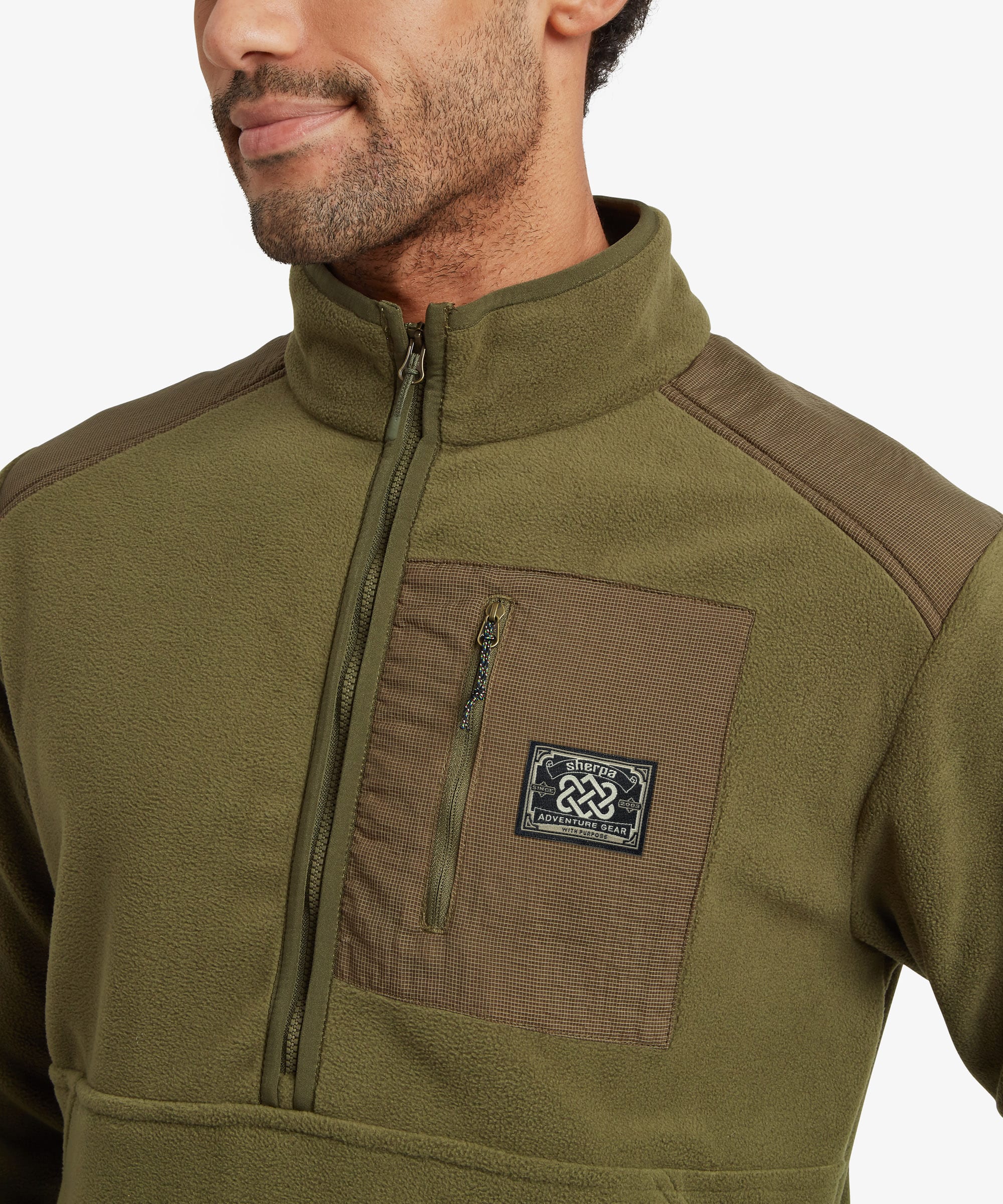 Close-up of a man wearing an olive green half zip fleece pullover with brown contrast panels on the chest and shoulders, featuring a zippered pocket and a small Sherpa Adventure Gear brand patch.