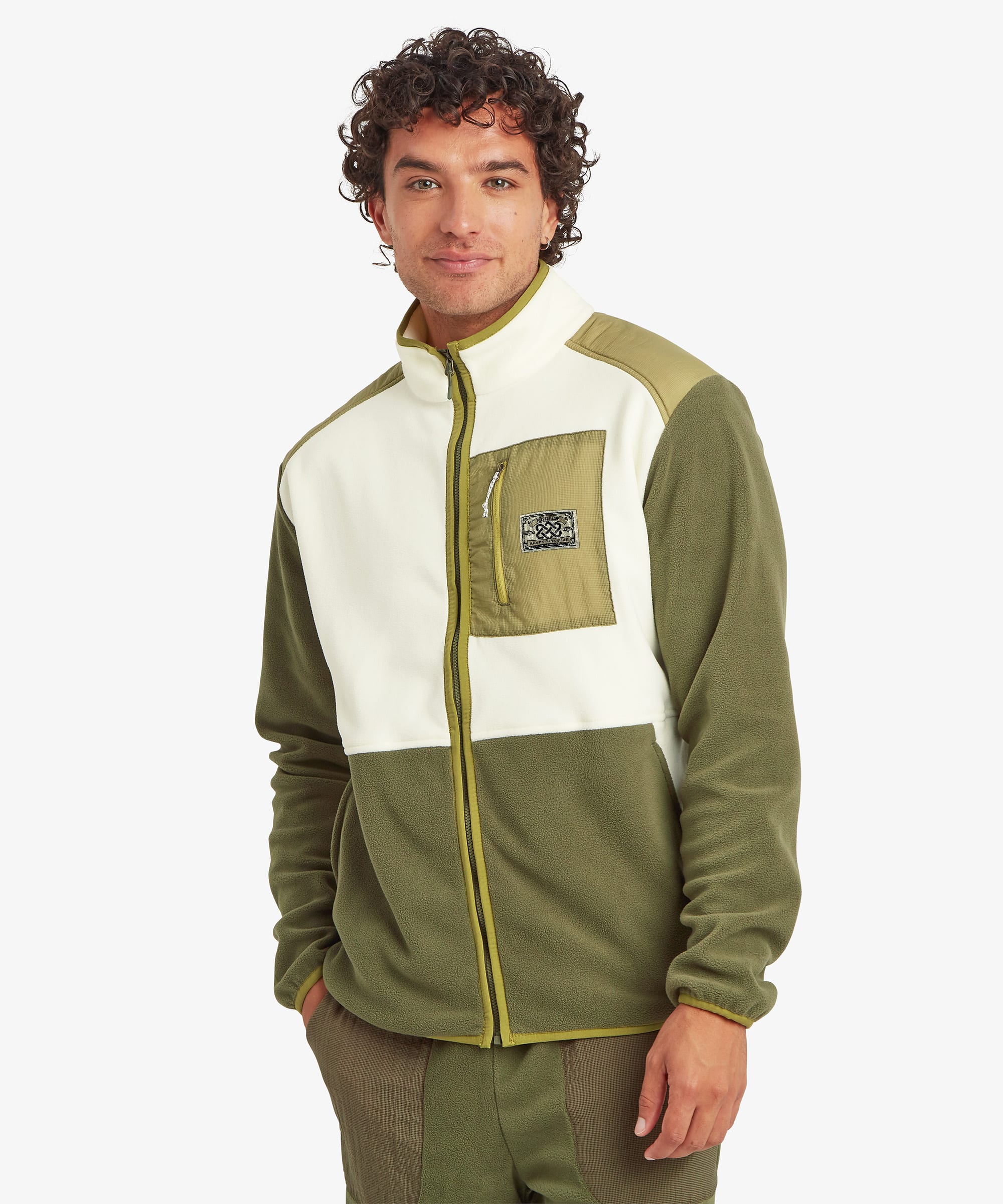 Front view of a male model wearing the Sherpa Sanu Full Zip in a cream and olive colour-block design with matching olive trousers. The fleece jacket features a high stand collar, full-length front zip, contrasting chest pocket with zip closure and Sherpa logo patch, elasticated cuffs and a relaxed fit.