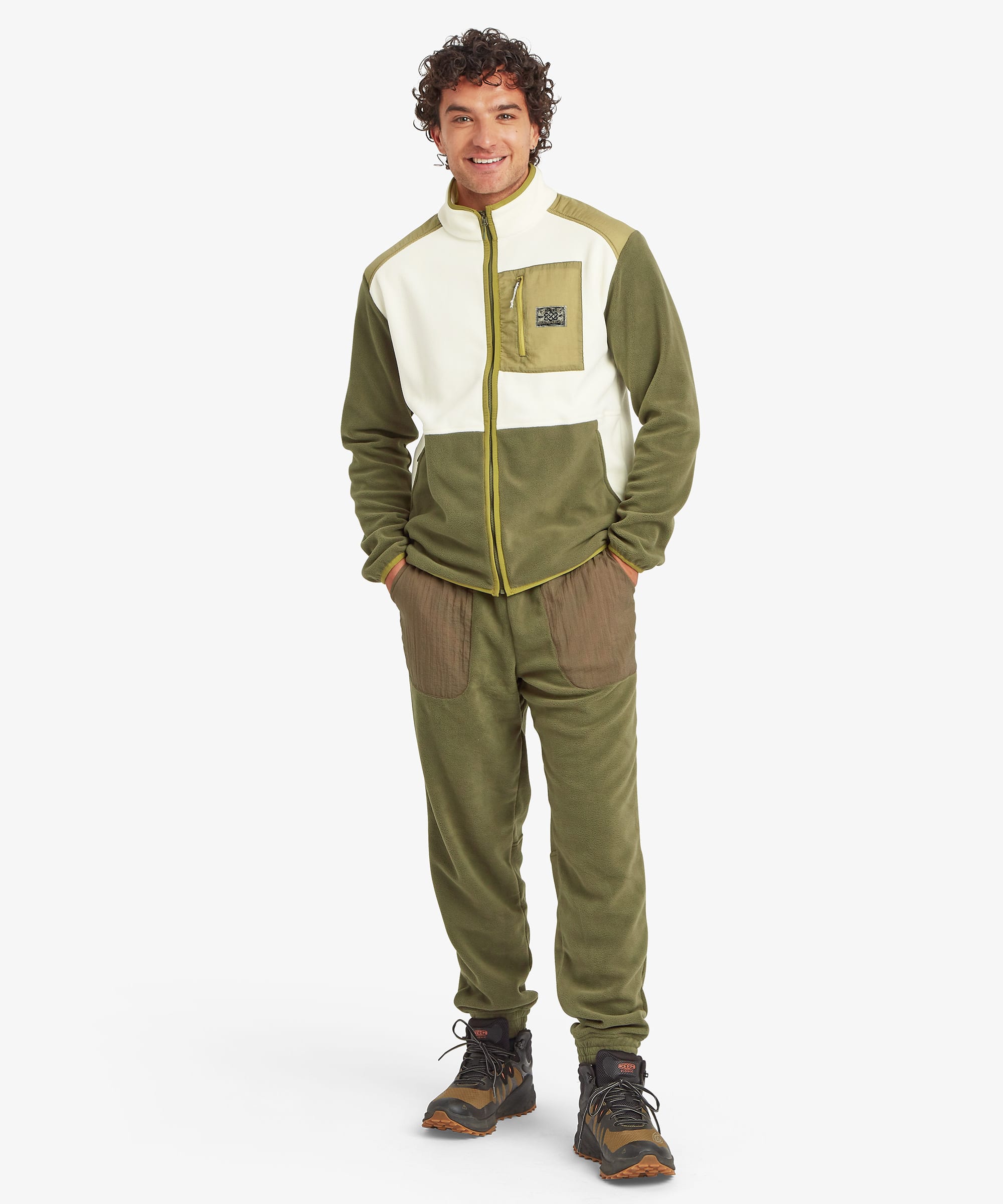 Full-length front view of a male model wearing the Sherpa Sanu Full Zip in cream and olive with matching olive trousers. The image highlights the colour-block fleece construction, stand collar, full zip fastening, zipped chest pocket, side pockets and relaxed outdoor fit.