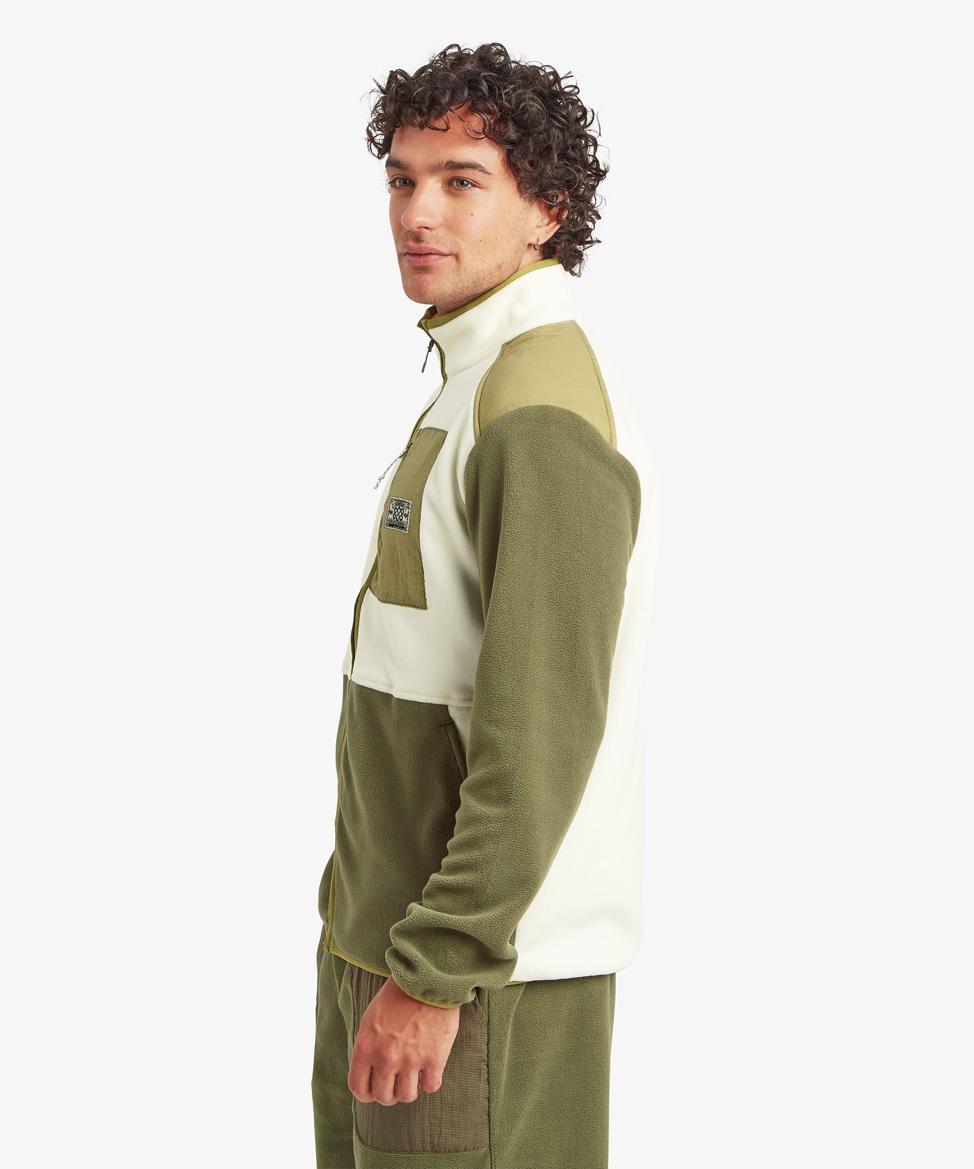 Side profile of a male model wearing the Sherpa Sanu Full Zip in cream and olive, showing the relaxed fit, high stand collar, long sleeves, colour-block panels and the soft fleece texture.