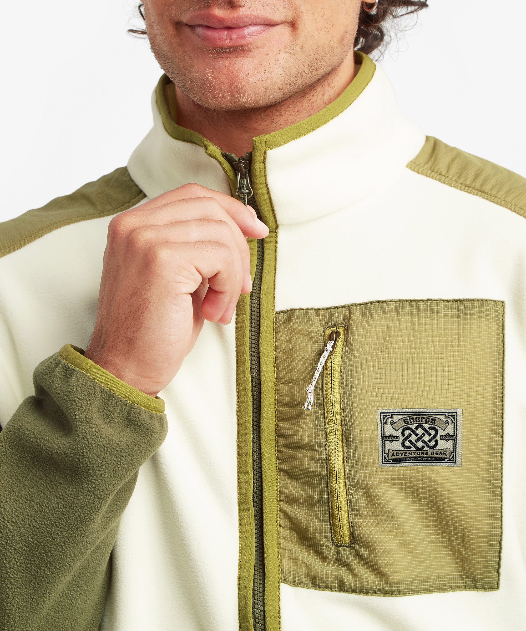 Close-up of a male model wearing the Sherpa Sanu Full Zip in cream and olive, highlighting the high stand collar, full front zip, contrasting zipped chest pocket, textured fleece fabric and woven Sherpa logo patch.