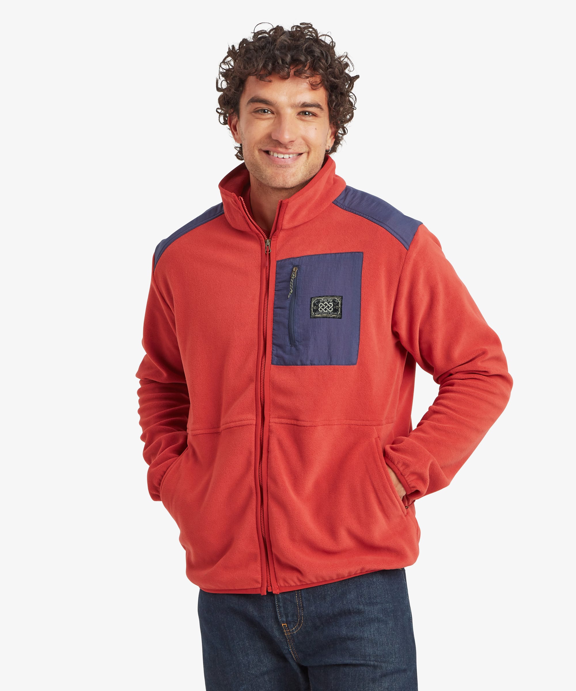 Front view of a smiling male model wearing the Sherpa Sanu Full Zip in red with contrasting navy shoulder panels and chest pocket, paired with dark blue jeans. The fleece jacket features a high stand collar, full-length front zip, zipped chest pocket with Sherpa logo patch, side hand pockets and a relaxed fit.