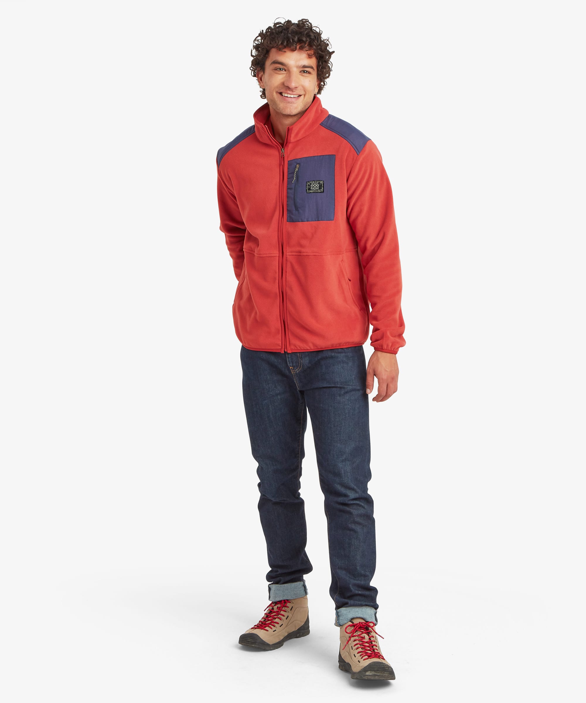 Full-length front view of a male model wearing the Sherpa Sanu Full Zip in red with navy colour-block details on the shoulders and chest pocket. The jacket has a stand collar, full zip fastening, side pockets, long sleeves and a relaxed everyday fit.