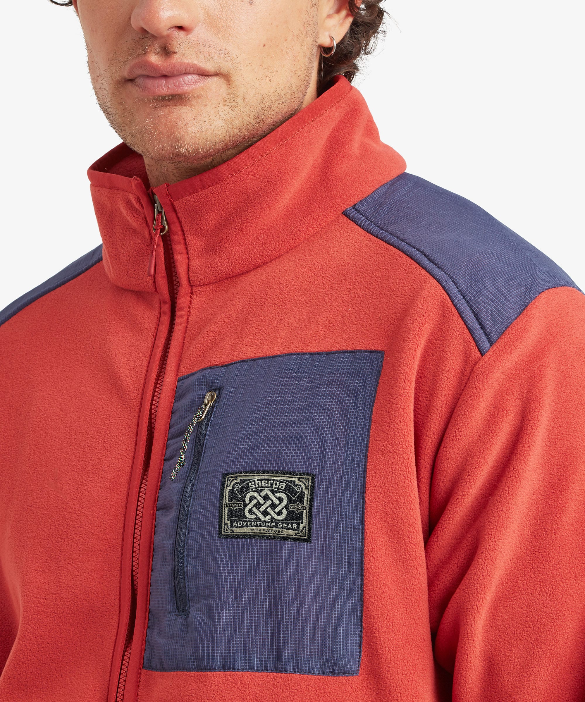 Close-up of a male model wearing the Sherpa Sanu Full Zip in red, highlighting the high stand collar, full front zip, contrasting navy zipped chest pocket, textured fleece fabric and woven Sherpa logo patch.