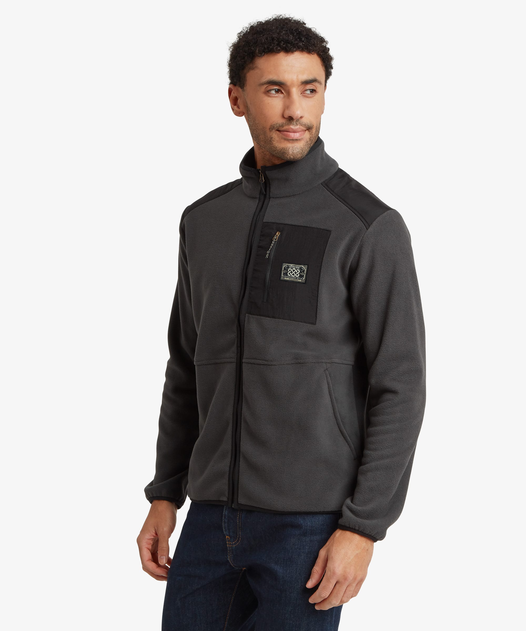 A man wearing a dark gray zip-up fleece jacket with black shoulder and chest panel details, a Sherpa Adventure Gear patch, paired with dark blue jeans, photographed against a light gray studio background.