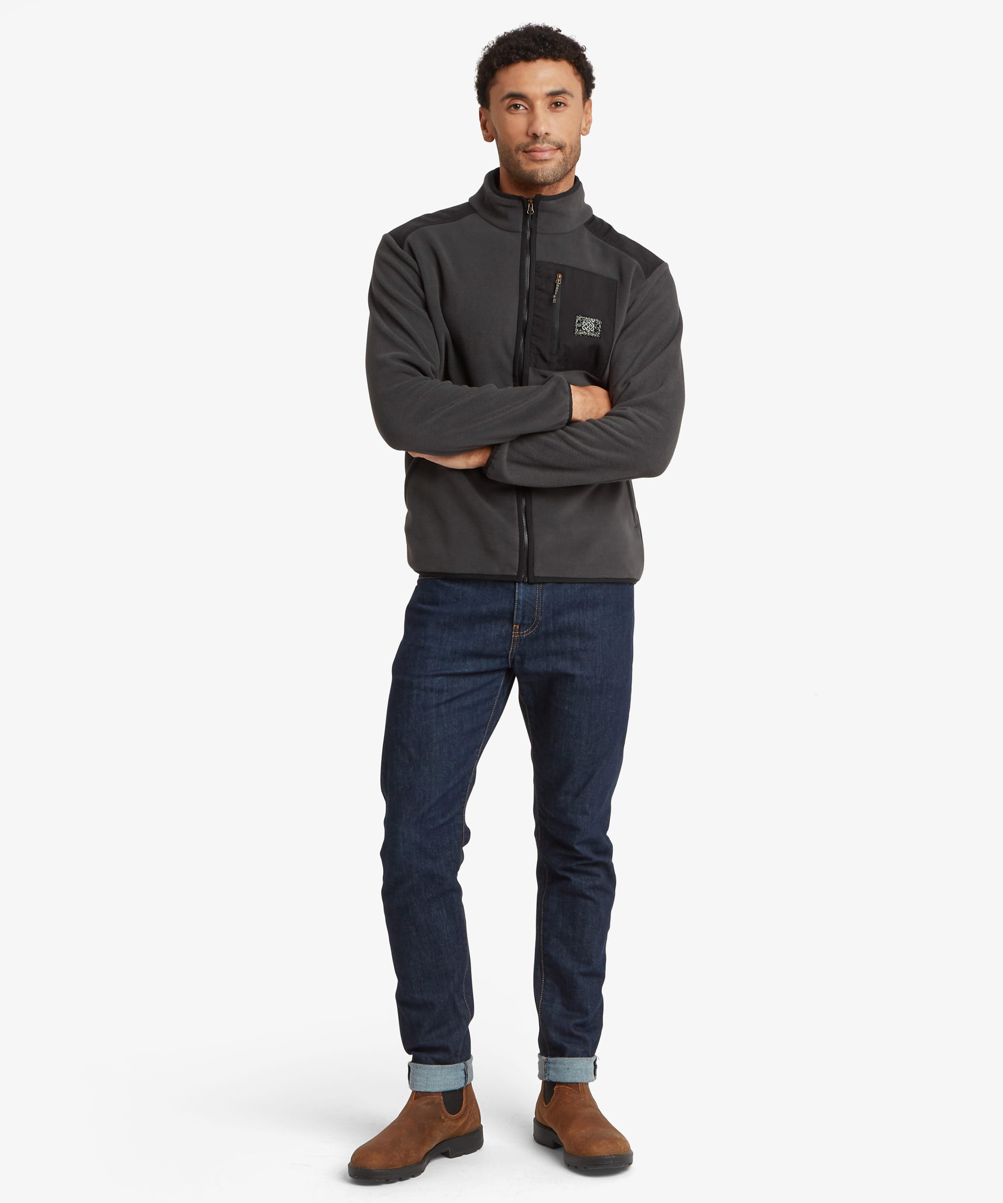 A man modeling a dark gray half-zip fleece jacket with a zippered chest pocket, a Sherpa Adventure Gear patch, paired with dark blue jeans and brown boots, standing against a light gray background.