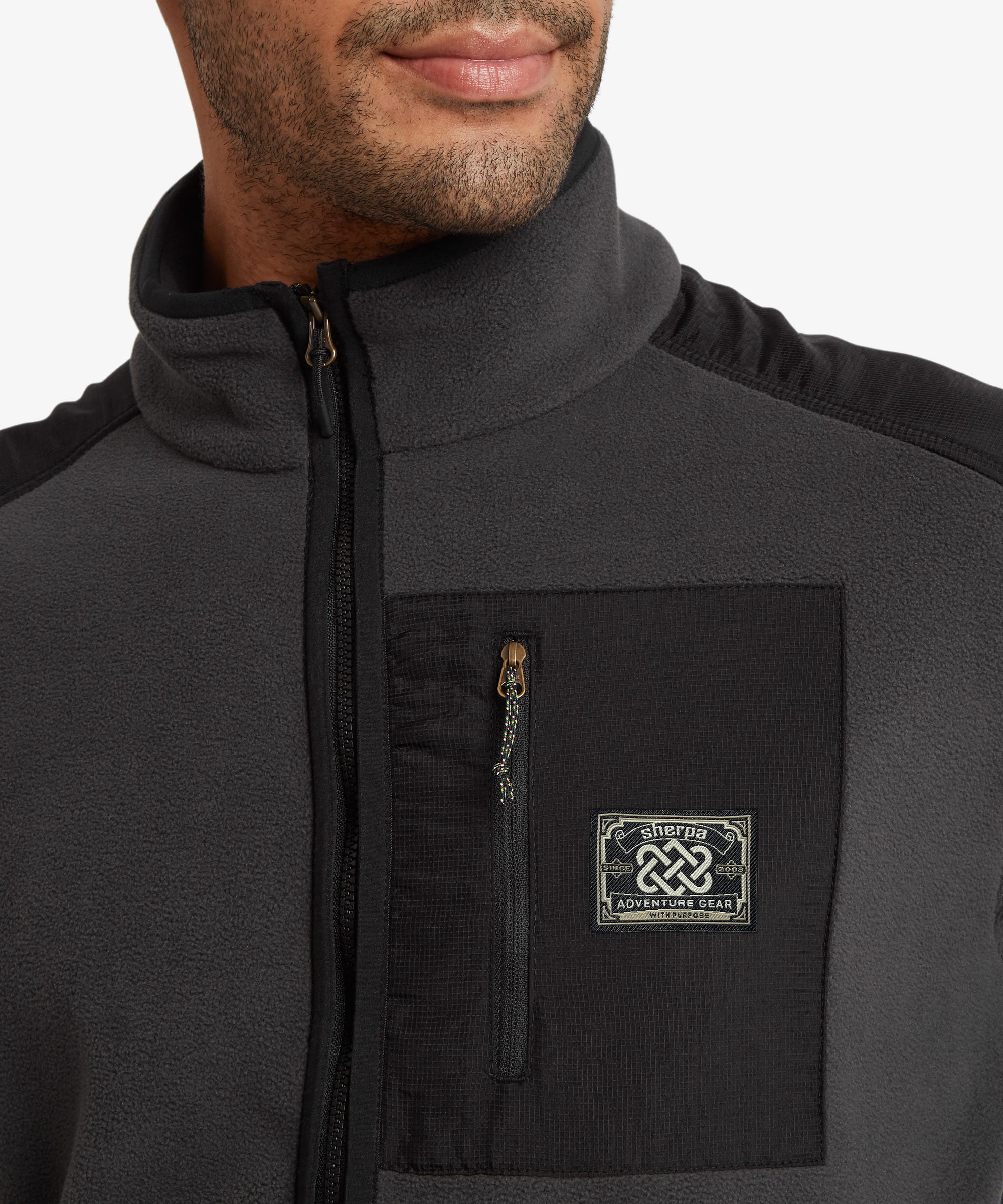 Close-up of a man wearing a dark gray and black fleece jacket with a high collar, a vertical chest zip pocket, and a square embroidered Sherpa Adventure Gearbrand patch on the chest pocket.