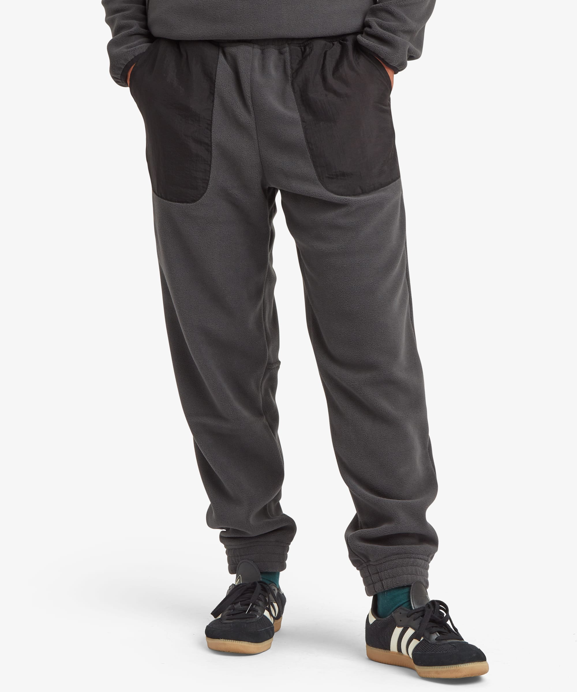Dark gray Sherpa Adventure Gear fleece jogger pants with contrasting black patch pockets, shown on a model from the waist down wearing black sneakers.