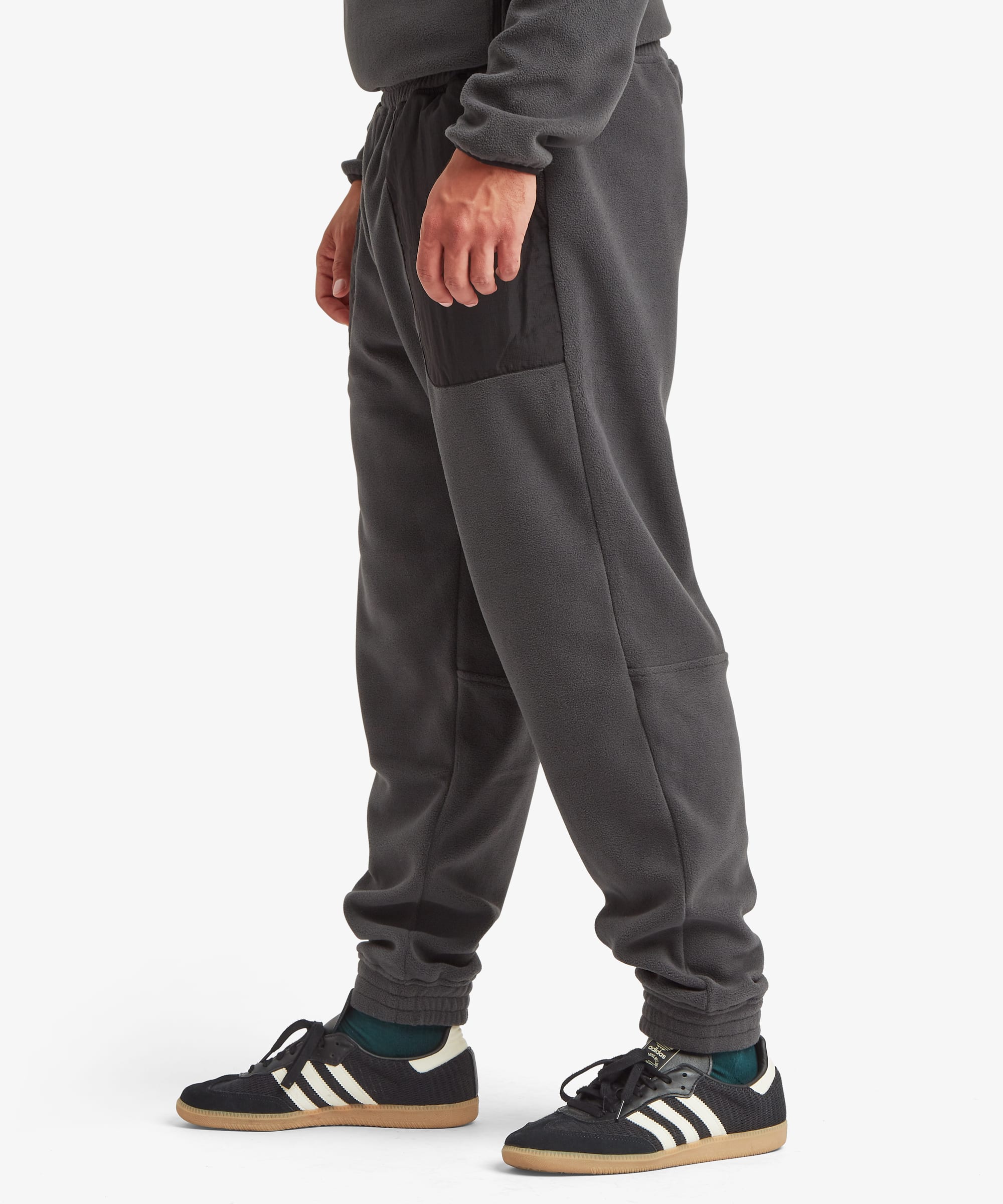 A person wearing dark gray jogger sweatpants with black side panel details, paired with black sneakers and teal socks, shown from the waist down against a light gray studio background.