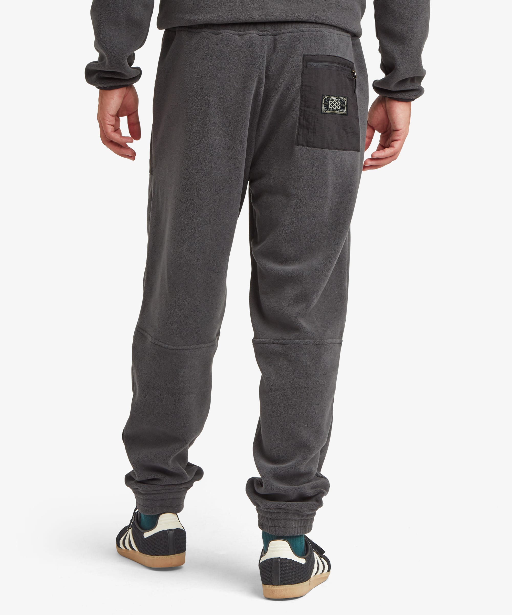 Rear view of a person wearing dark gray sweatpants with a back pocket and logo patch, paired with black and green sneakers, against a light gray studio background. The sweatpants feature a black patch with an endless knot and writing that reads "Sherpa Adventure Gear".