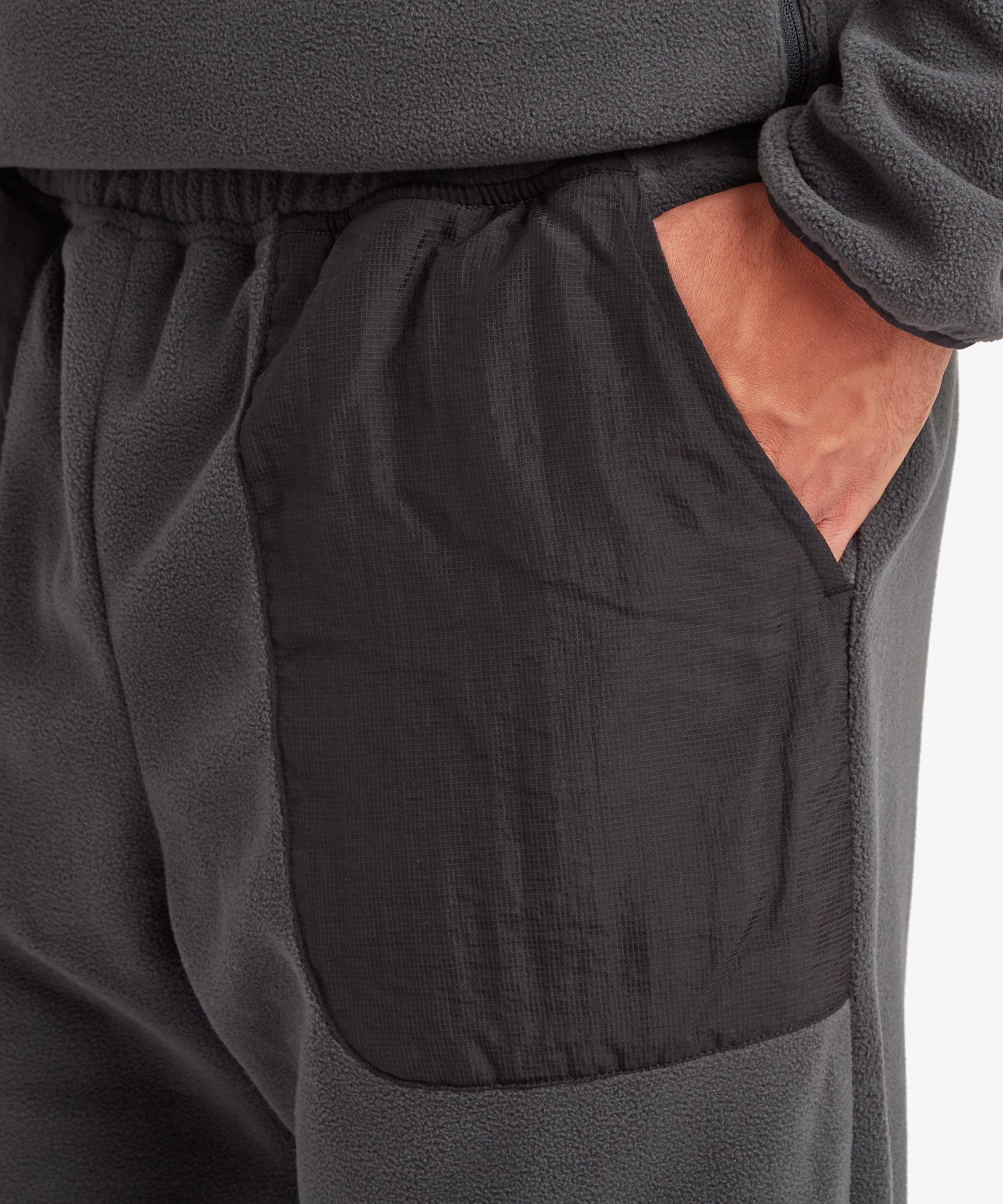 Close-up of a person wearing dark charcoal fleece pants, showing a side pocket with a hand in the pocket.The pocket is made of a black rip-stop fabric.