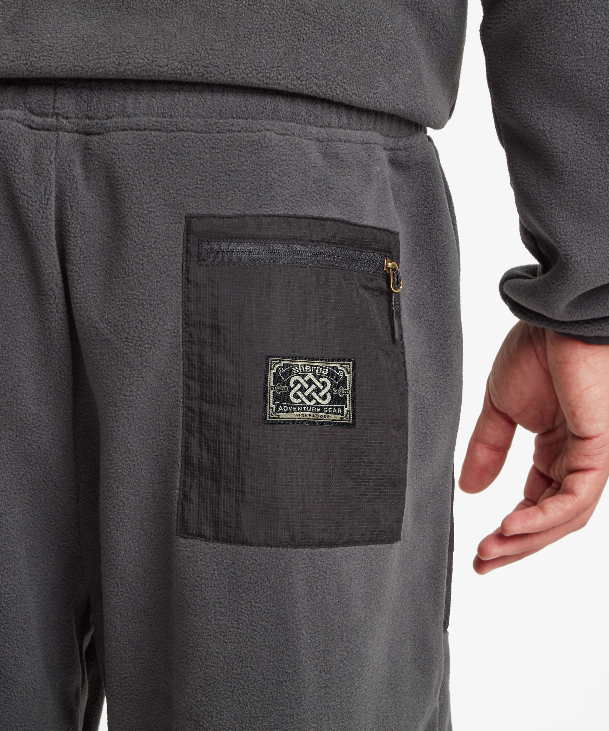 Close-up of dark gray fleece sweatpants showing a side pocket with a zip closure and a square brand patch that reads "Sherpa Adventure Gear" and "Tarcho Collection" A person's hand is visible at the side.