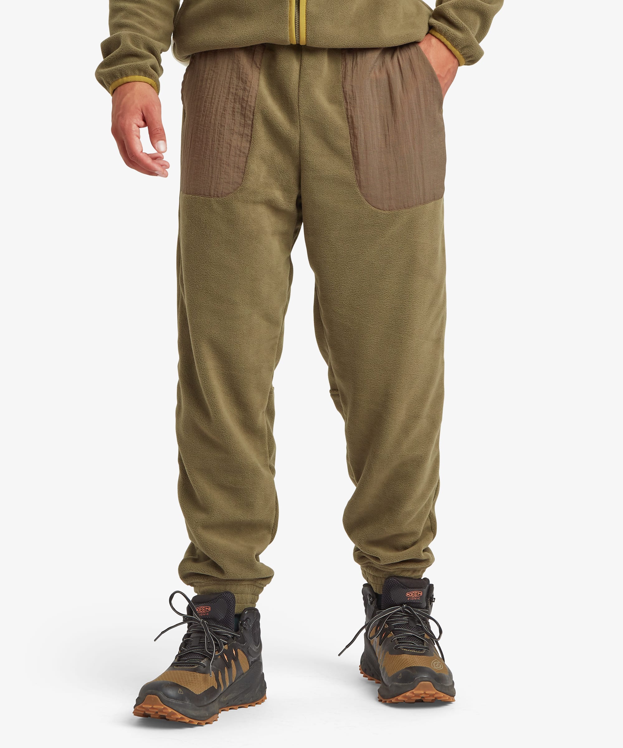 Front view of olive green jogger pants with contrasting brown patch pockets on the thighs, elastic cuffs at the ankles, and a relaxed fit. Styled with hiking boots.