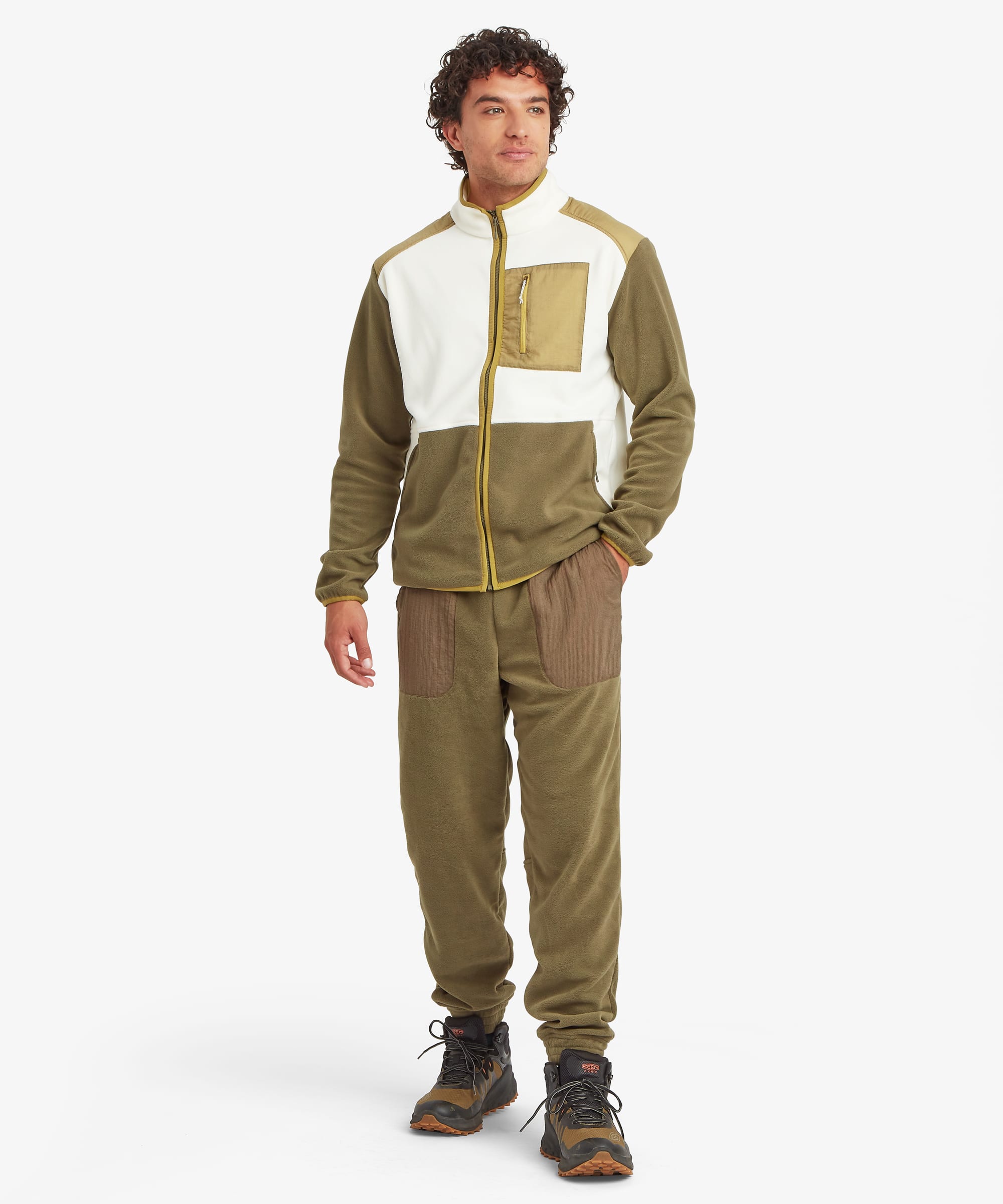 Full-body front view of a model wearing olive green jogger pants with oversized contrast thigh patch pockets, elastic waistband, and cuffed hems. Coordinated with a matching color-block fleece jacket.