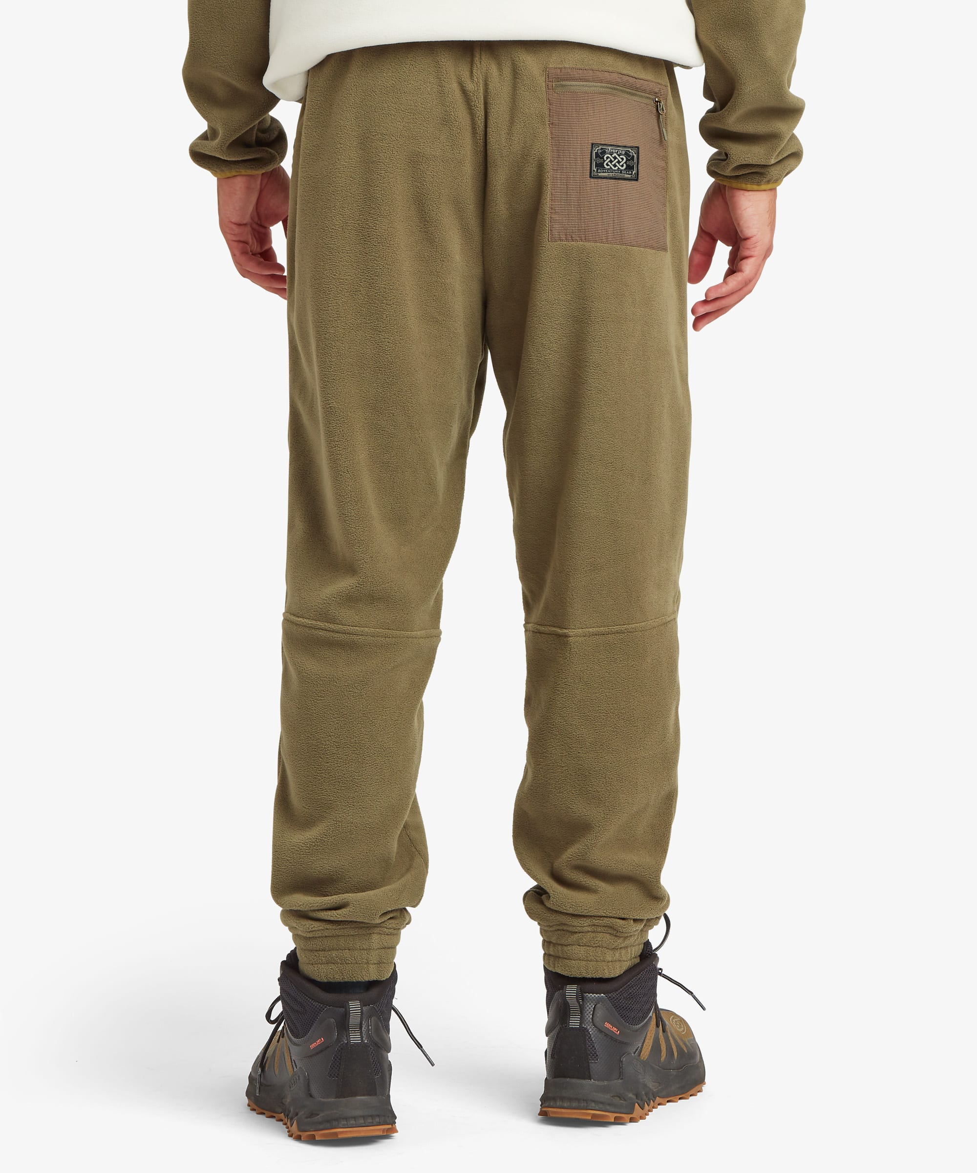 Rear view of a person wearing olive green fleece jogger pants with a brown back pocket patch and black hiking boots, against a light gray studio background.