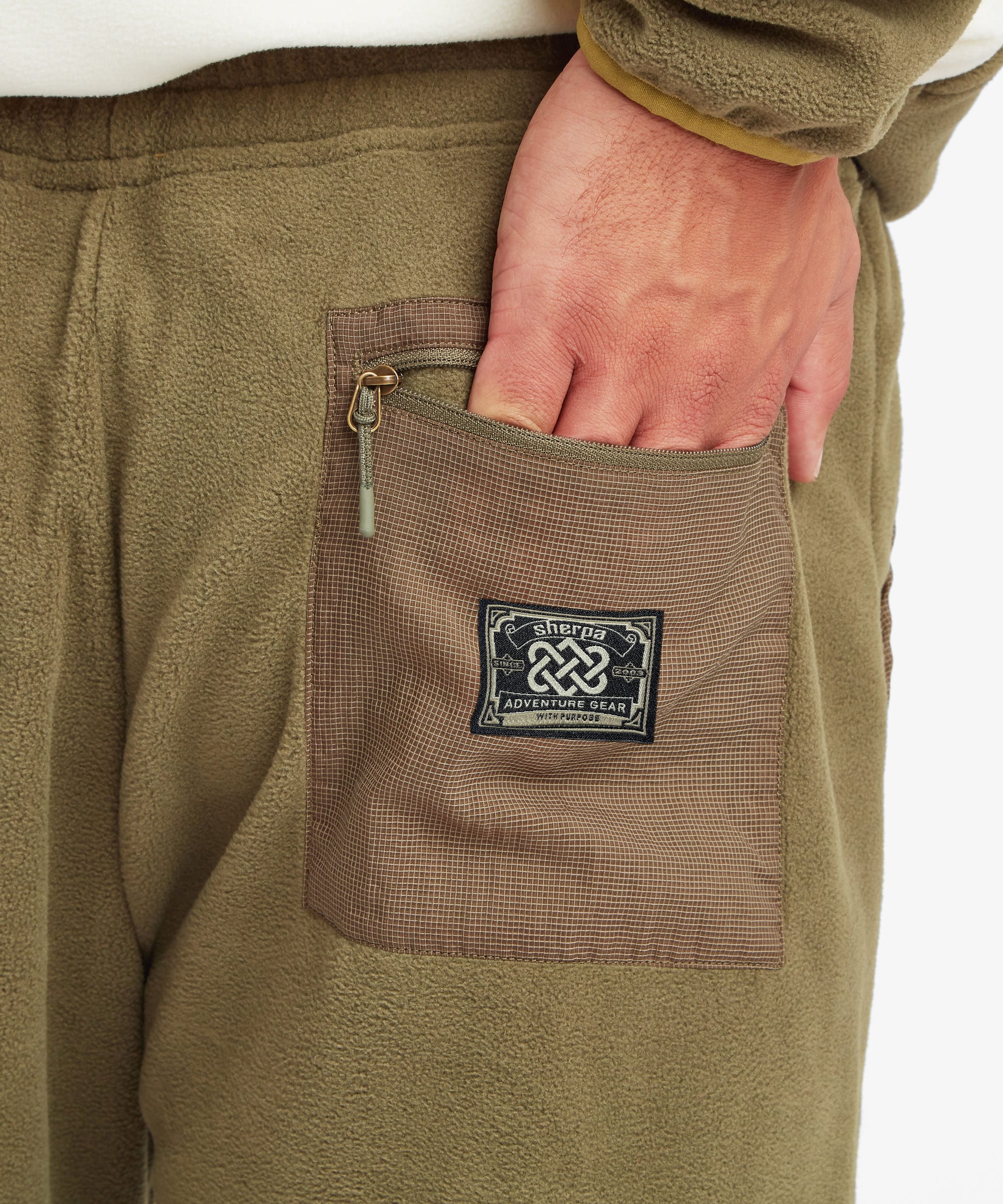 Close-up of a person wearing olive brown fleece pants with a brown utility pocket and a black Sherpa Adventure Gear brand patch. The man is reaching into the pocket with his hand.