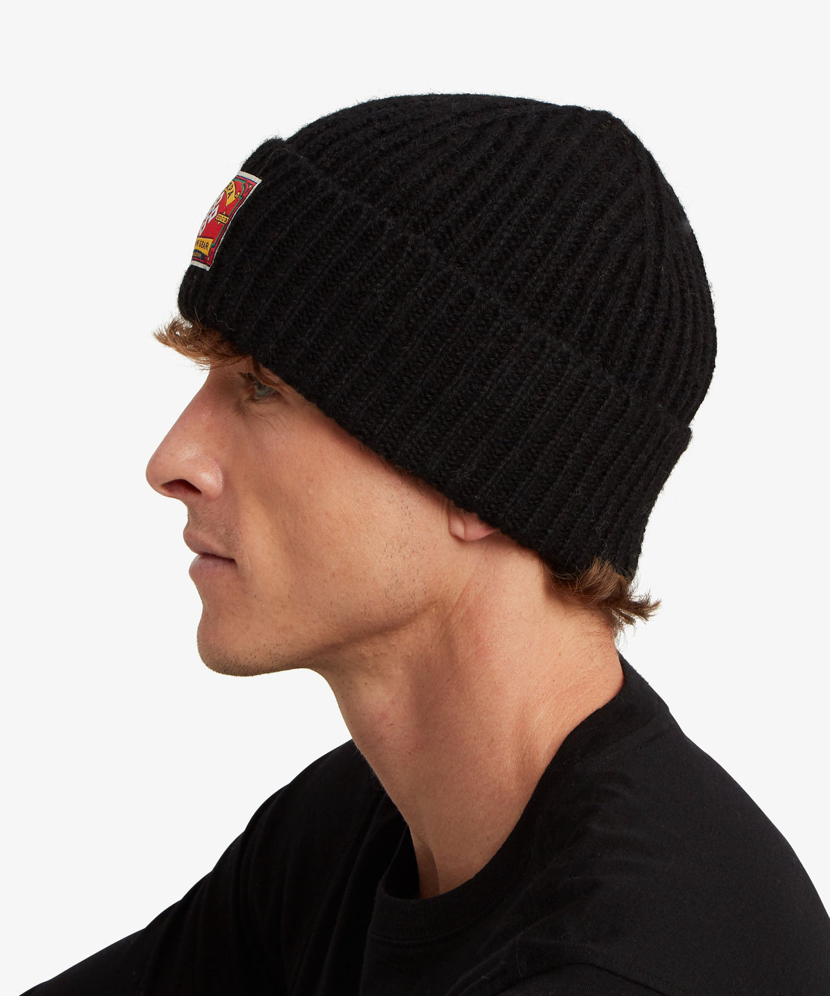 A man in profile wearing a black ribbed knit beanie with a small red.