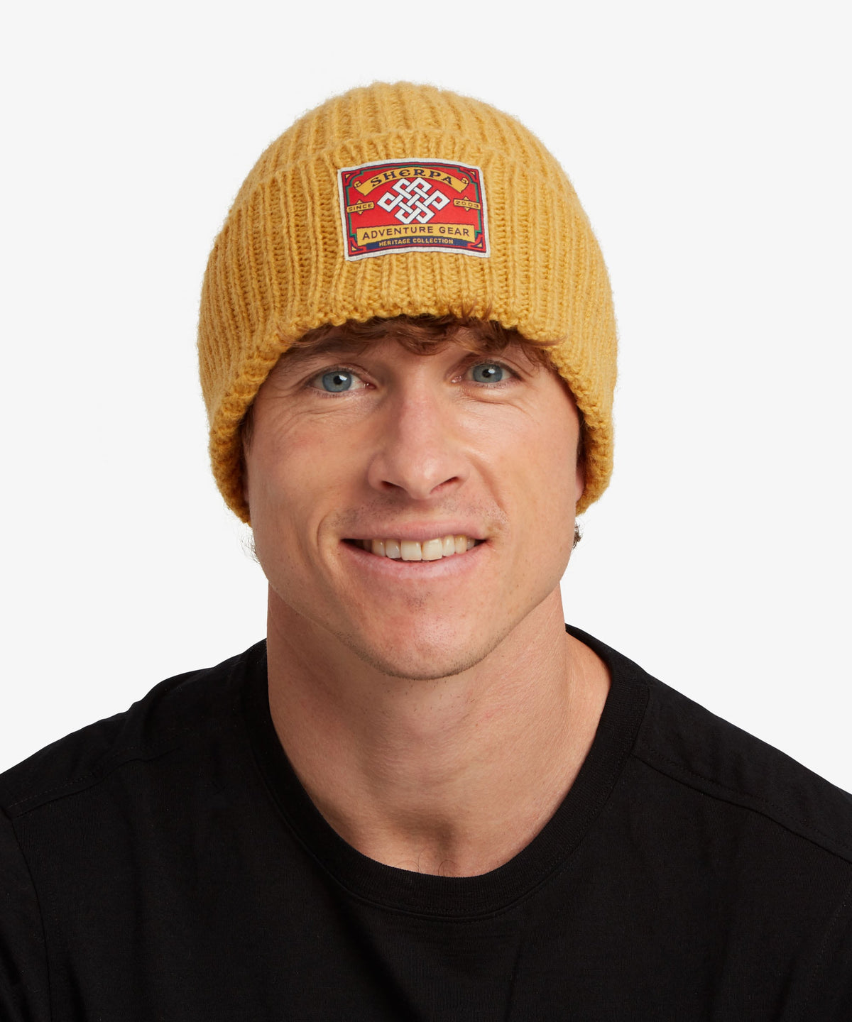 A smiling man wearing a mustard yellow ribbed knit beanie with a rectangular 'Sherpa Adventure.