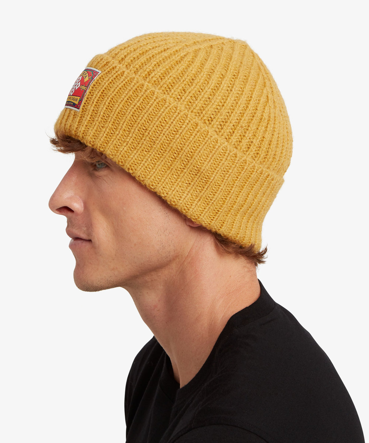 A man in profile wearing a mustard yellow ribbed knit beanie with a small embroidered patch.