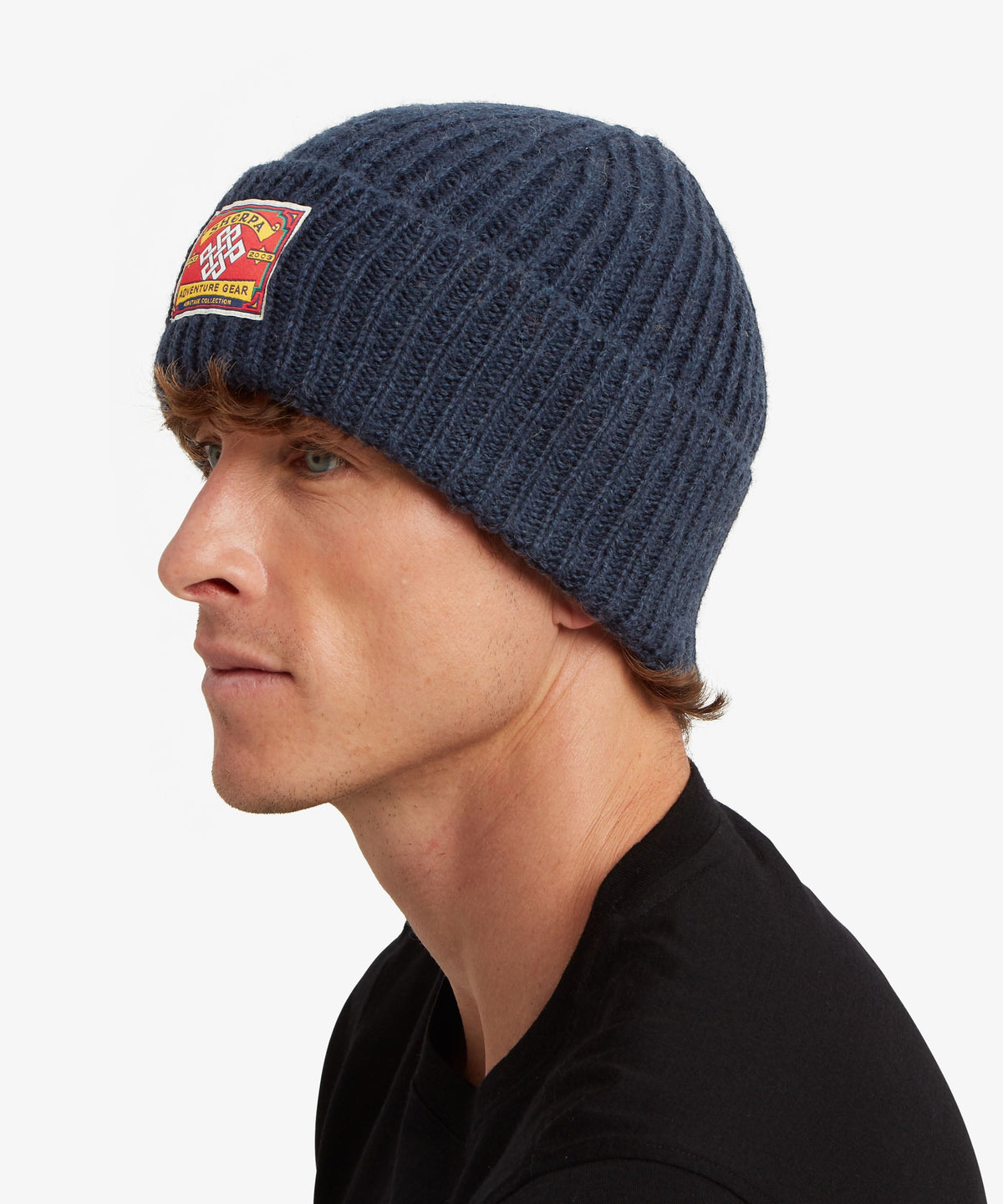 A man in profile wearing a navy blue ribbed knit beanie with a red and yellow patch on the side.