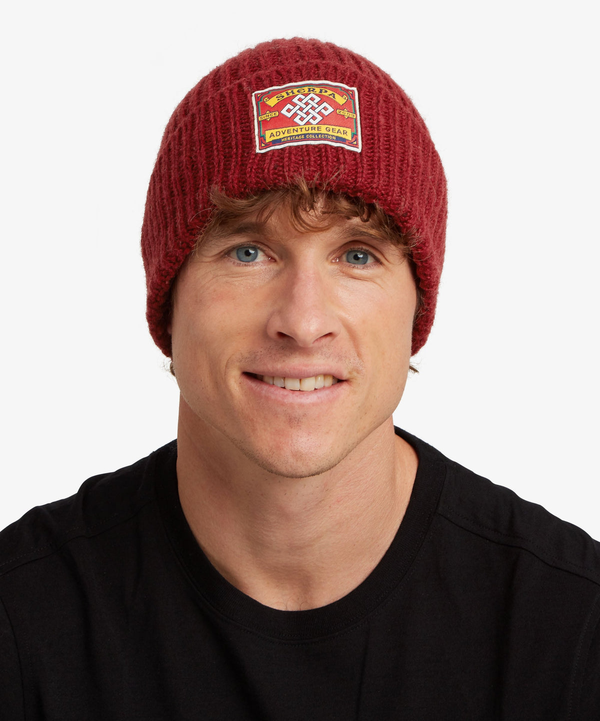 A smiling man wearing a dark red ribbed knit beanie with a rectangular embroidered patch.