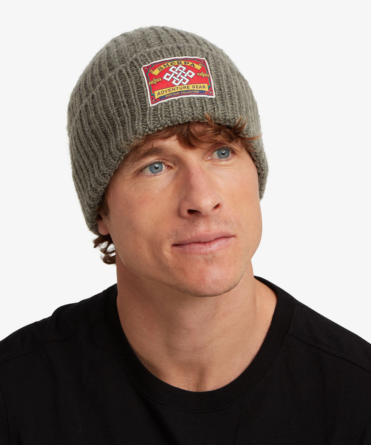 A man wearing a ribbed muted olive beanie with a red and yellow 'Sherpa Adventure Gear' patch.