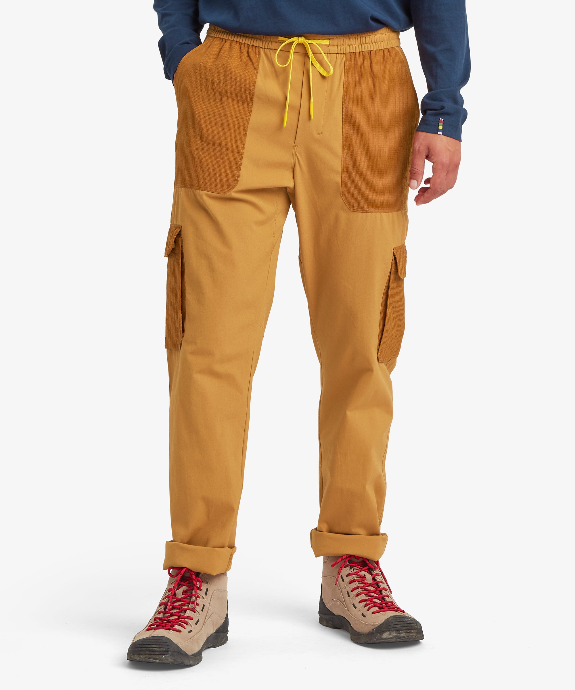 A front view of mustard-yellow cargo trousers with textured ripstop patch pockets, an elastic waistband with a yellow drawstring, and cuffed hems. Styled with tan hiking boots.